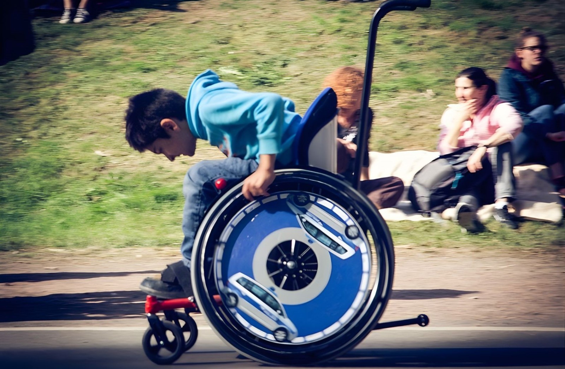 Ein jugendlicher selbstfahrender Rollstuhlfahrer fährt auf einem asphaltierten Weg von. Er ist von der Seite zu sehen. Das Foto hat eine deutliche Bewegungsunschärfe, die zeigt, dass der Junge schnell unterwegs ist.