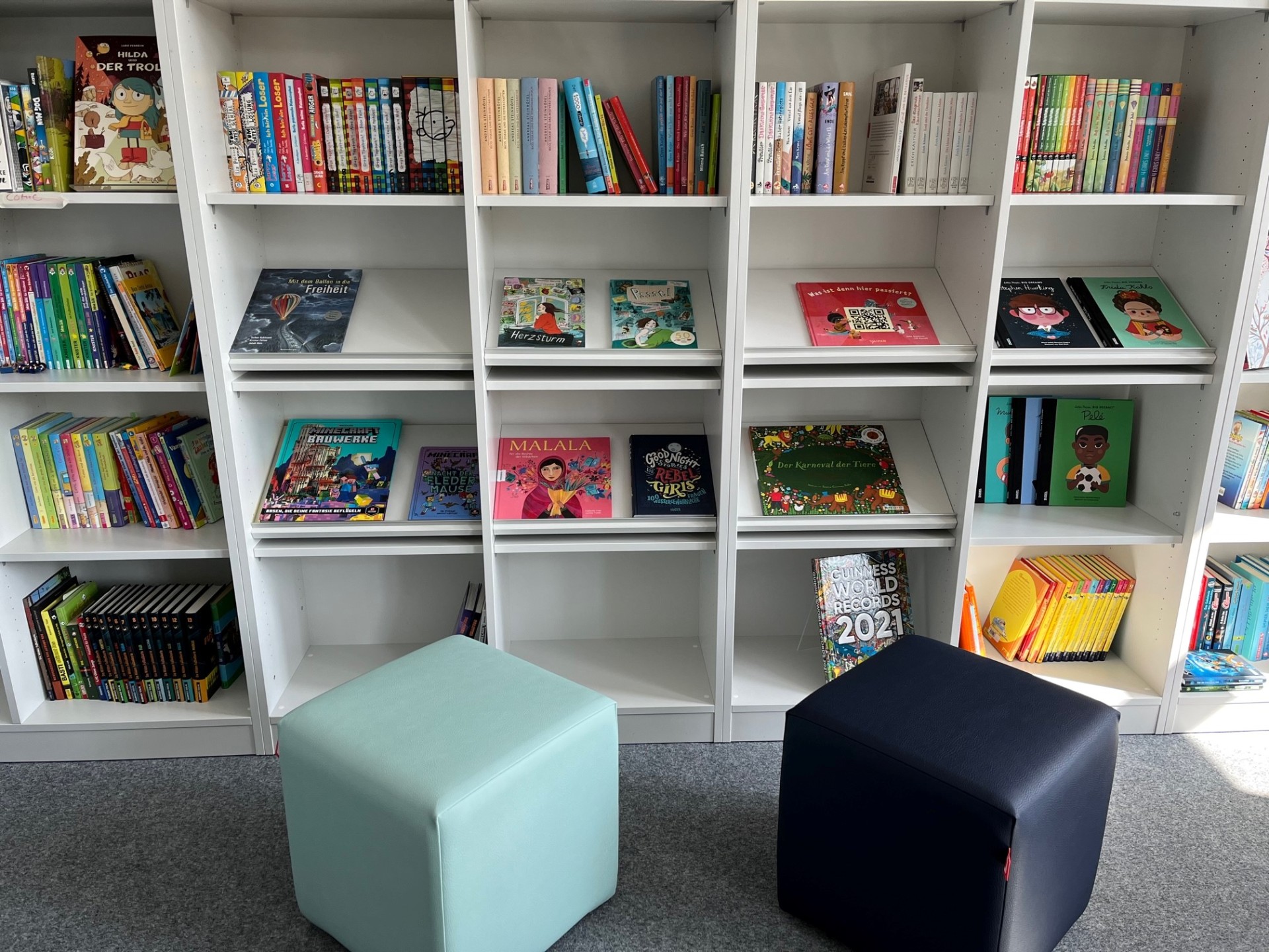 Ein helles, weißes Bücherregal in einer Bibliothek oder Schule, das mit zahlreichen Kinder- und Jugendbüchern gefüllt ist. Die Bücher sind teils stehend mit dem Rücken nach vorne, teils frontal auf Präsentationsleisten ausgestellt. Vor dem Regal stehen zwei quadratische Sitzhocker in Hellblau und Dunkelblau auf einem grauen Teppichboden.