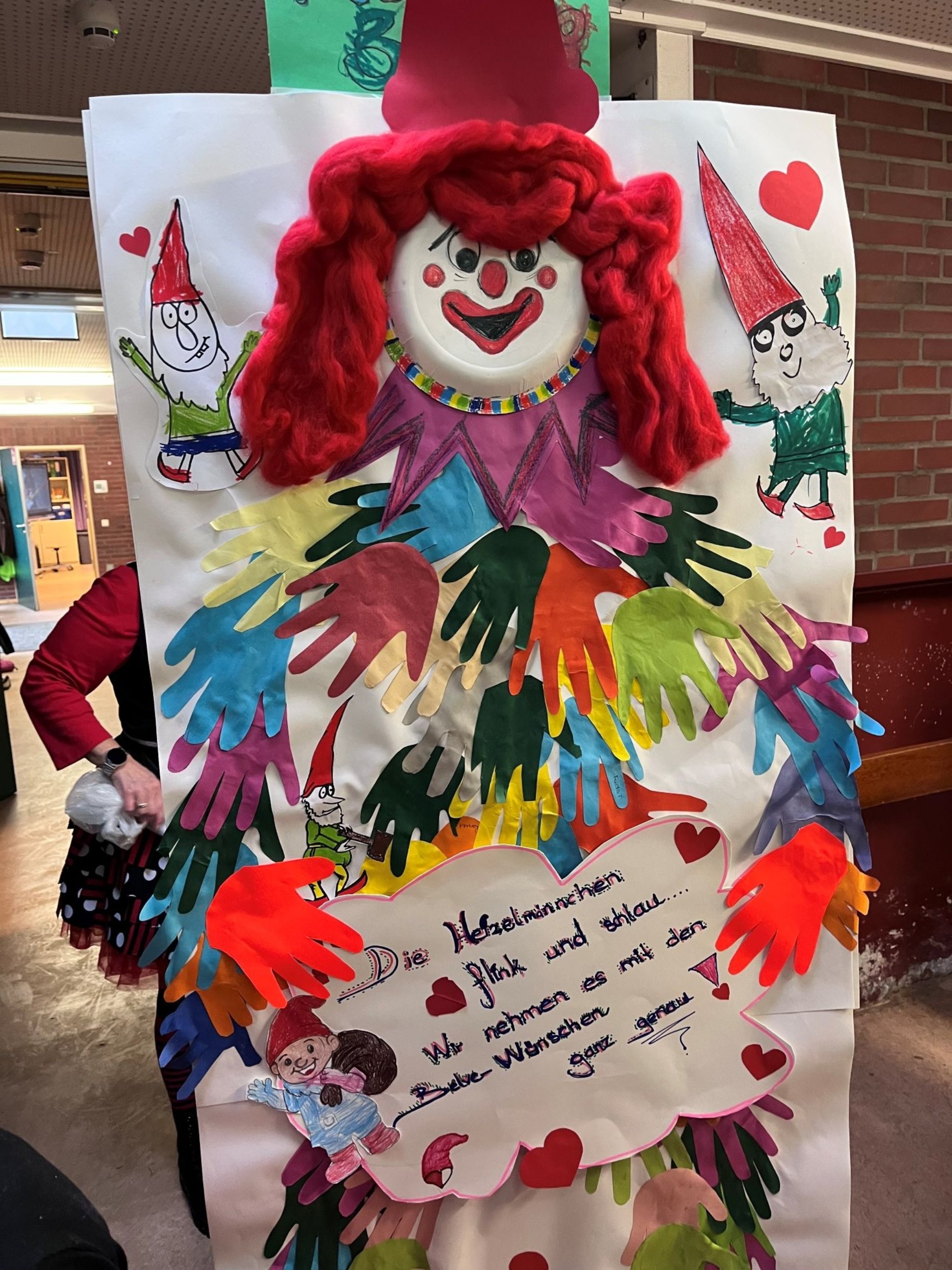 Ein großer, bunter Pappclown mit roter Wollperücke steht in einem Gang. Sein Körper besteht aus farbigen Handabdrücken. Kleine Wichtel und eine Texttafel mit „Die Heinzelmännchen“ umgeben ihn.