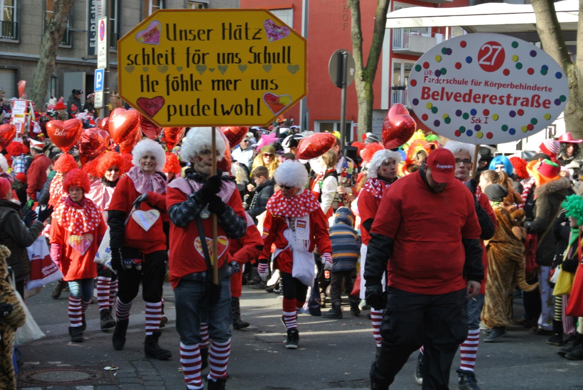 Eine als Clowns verkleidete Gruppe läuft durch eine Menschenmenge. Zwei Personen vorne tragen Schilder mit den Aufschriften: "27, LVR-Förderschule für Körperbehinderte Belvederestraße, Sek I" und "Unser Hätz schleit för uns Schull. He föhle mer uns pudelwohl." 