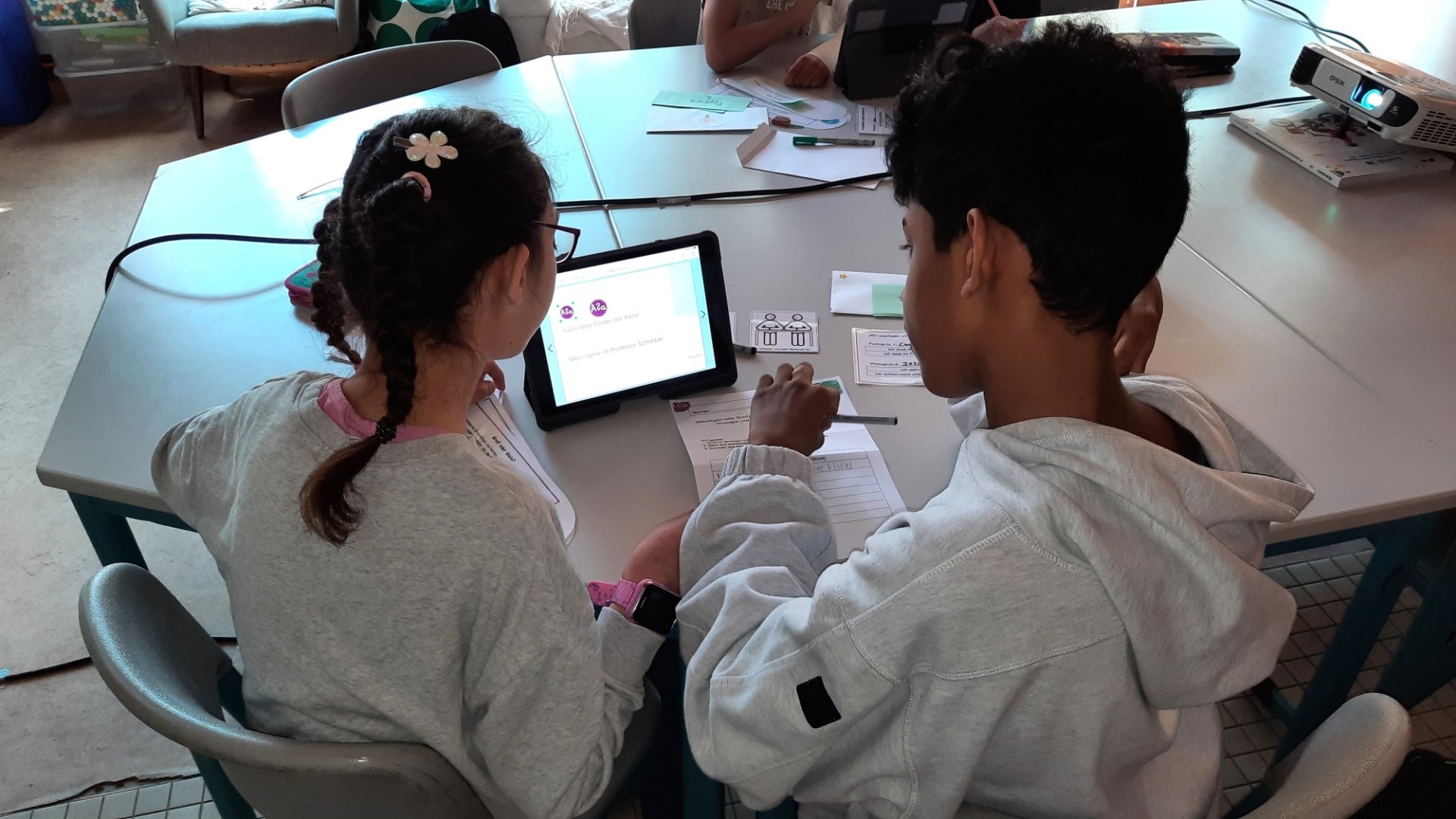 Zwei Schulkinder sitzen gemeinsam an einem Tisch und arbeiten konzentriert mit einem Tablet und Arbeitsblättern.