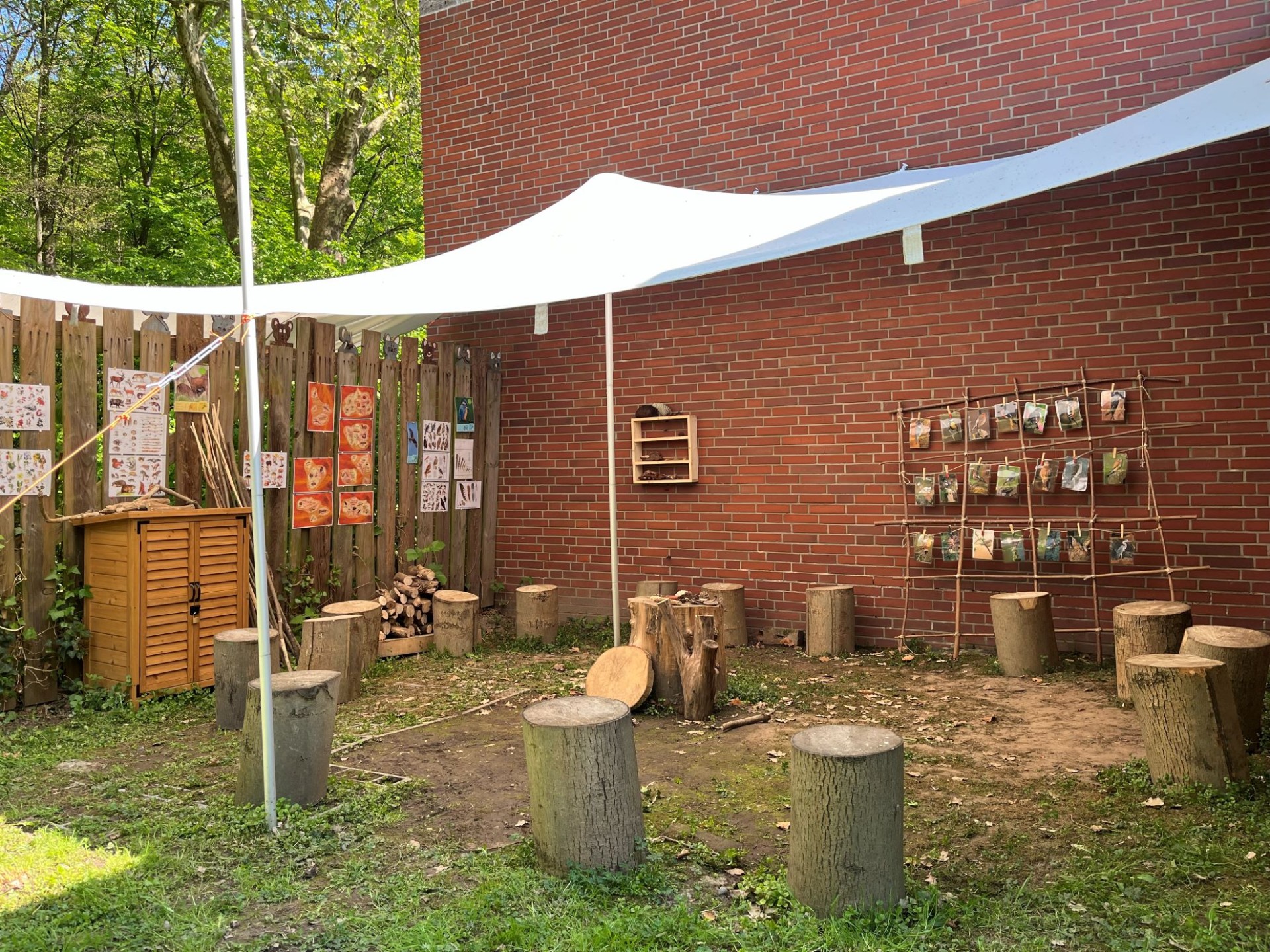 Ein Außenklassenzimmer auf einem Waldgrundstück mit Baumstämmen als Sitzgelegenheiten unter einem weißen Sonnensegel. An der Holzwand und dem Backsteingebäude hängen Bilder.