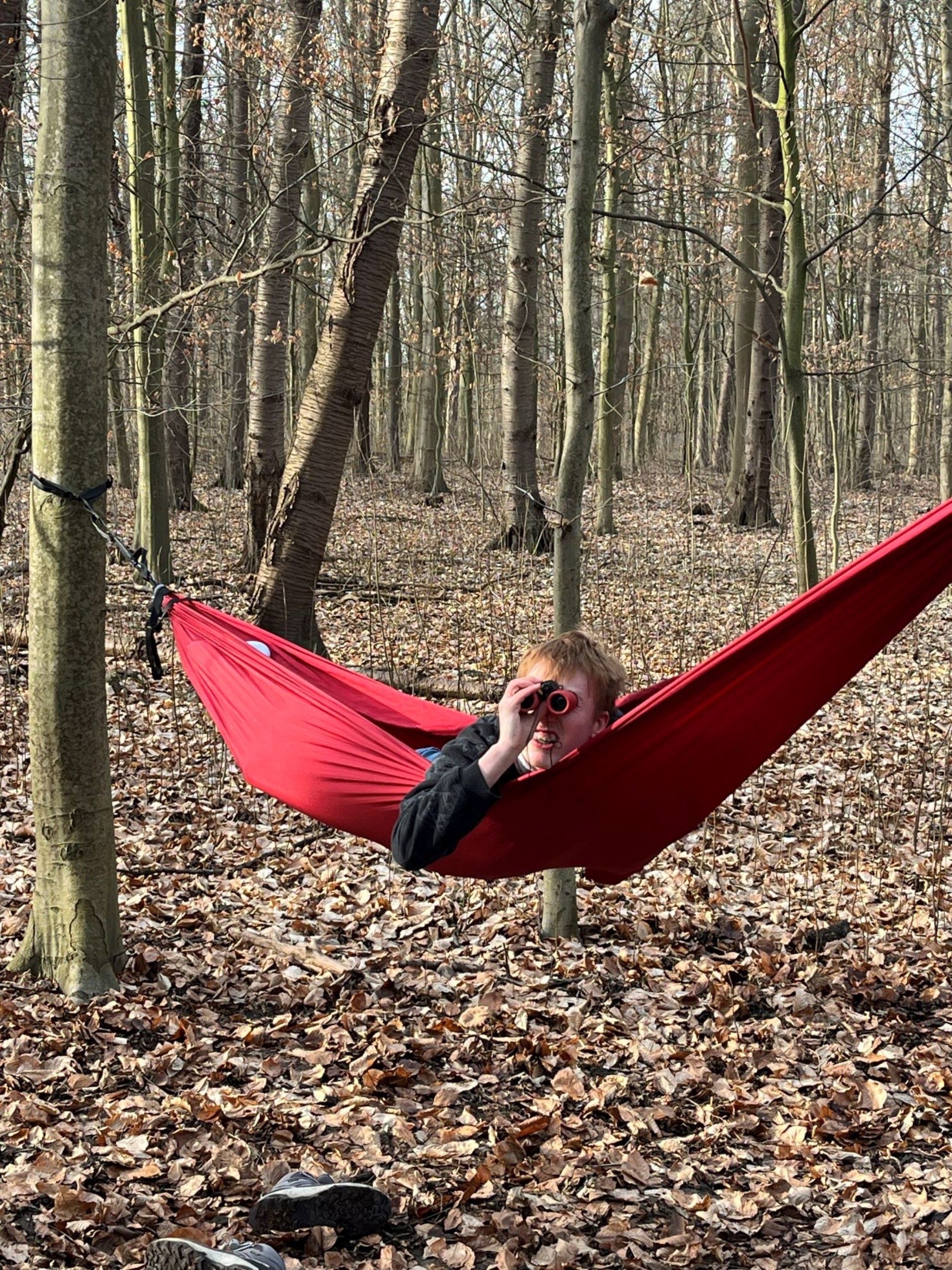 Ein Jugendlicher mit rötlichem Haar liegt in einer roten Hängematte im Wald und schaut durch ein Fernglas direkt in die Kamera. Der Boden ist mit braunem Laub bedeckt.