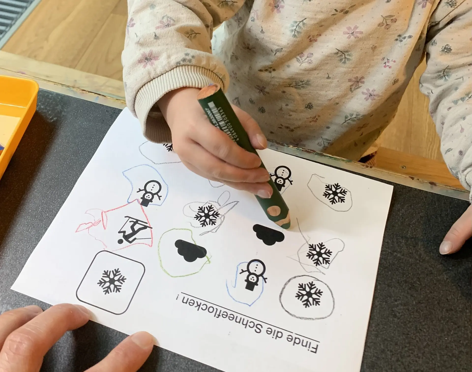 Ein Kind hält einen Buntstift in der rechten Hand und zeichnet auf ein Arbeitsblatt mit der Überschrift "Finde die Schneeflocken!". Die Aufgabe beinhaltet, Schneeflocken-, Schneemann- und Wolken-Symbole zu unterscheiden.