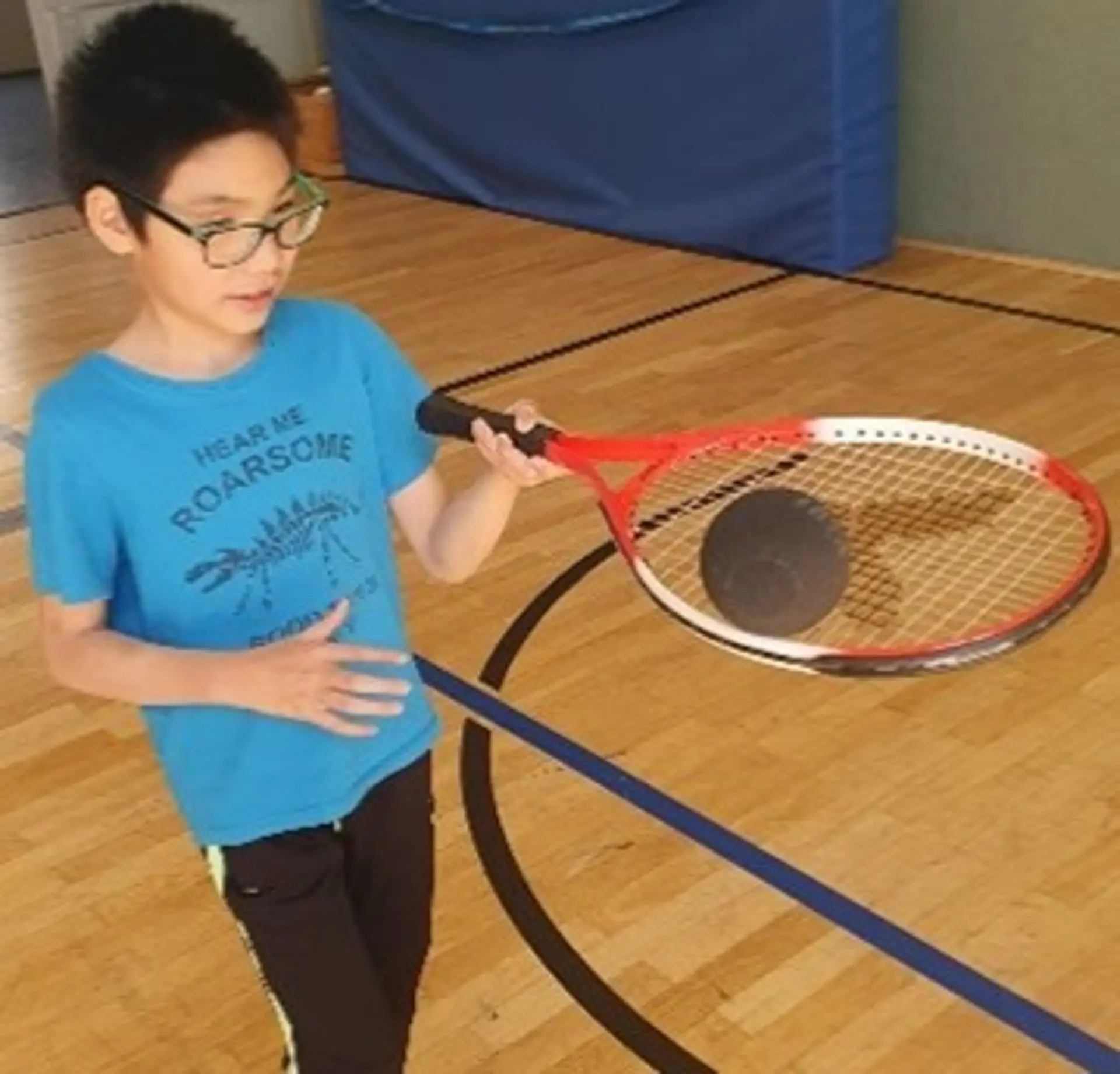 Ein Schüler mit Brille balanciert auf einem Tennisschläger einen Ball für Blinden-Tennis. Er ist in einer Sporthalle.