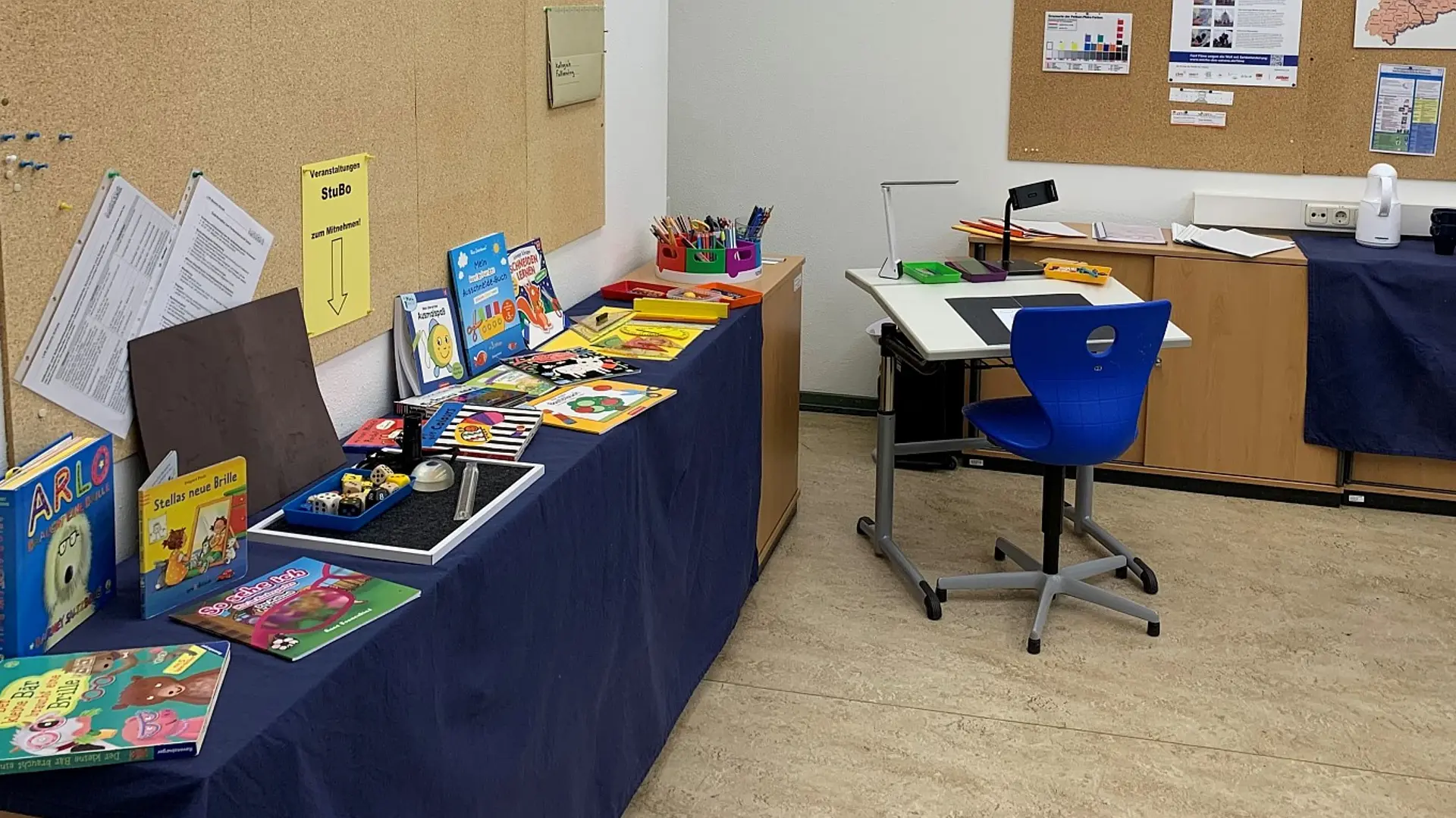 Eine Ecke eines Klassenzimmers, wo ein langer Tisch mit einer dicken Tischdecke darauf steht. Auf dem Tisch sind verschiedene Bücher und Utensilien für sehbeeinträchtige Kinder ausgestellt.