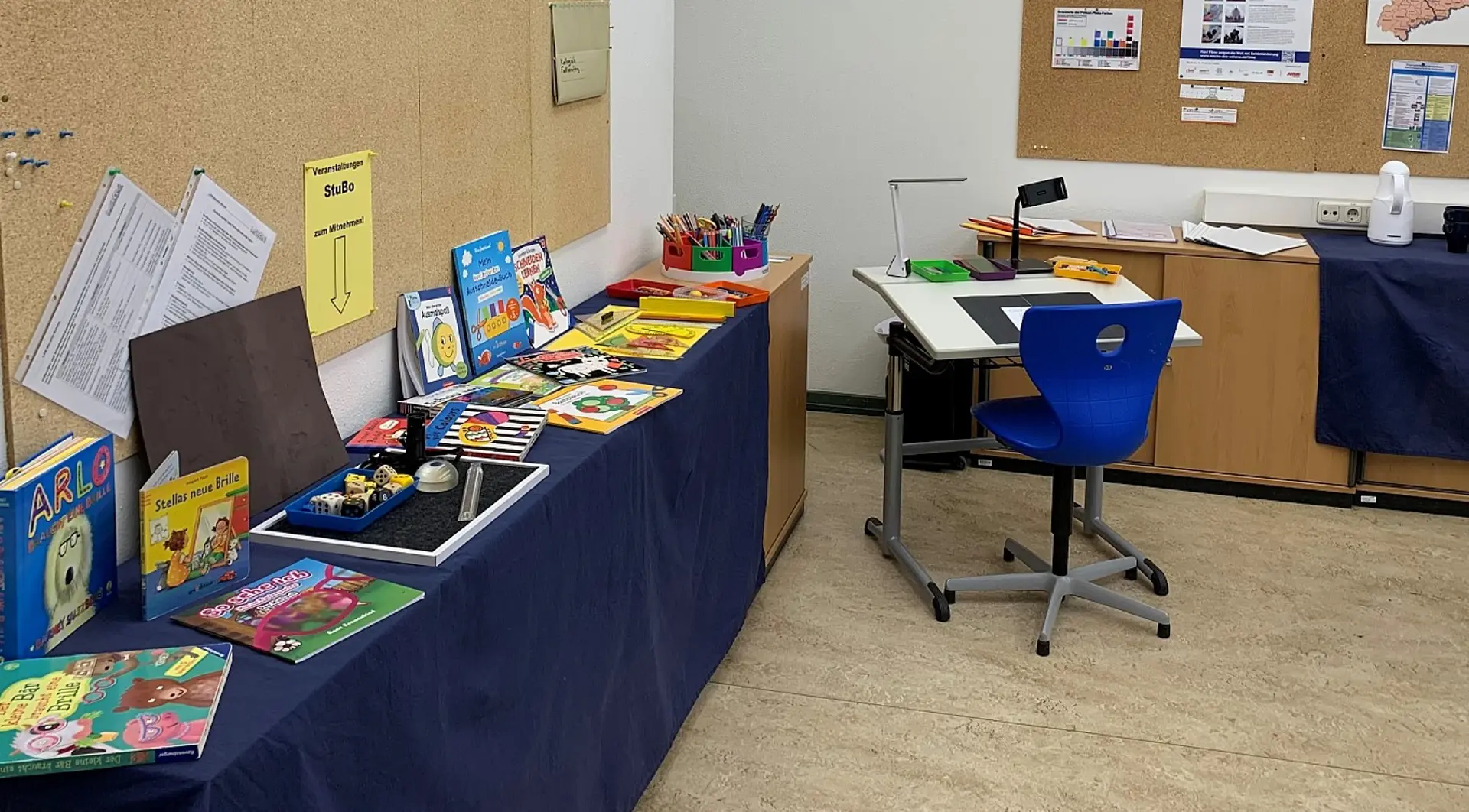 Eine Ecke eines Klassenzimmers, wo ein langer Tisch mit einer dicken Tischdecke darauf steht. Auf dem Tisch sind verschiedene Bücher und Utensilien für sehbeeinträchtige Kinder ausgestellt.