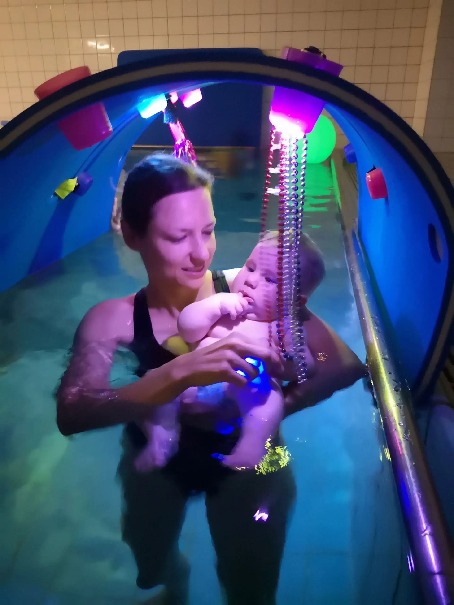 Eine Frau hält ein Baby, es liegt entspannt mit dem Rücken auf ihrem Arm. Sie befinden sich im Schwimmbecken, tragen Badekleidung und gehen durch einen bunt ausgeleuchteten Tunnel