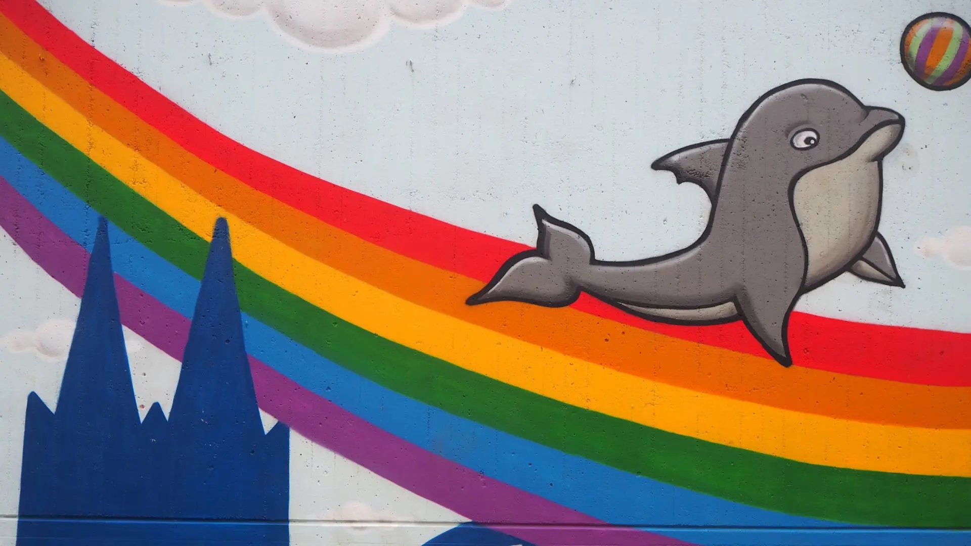 Ein Teil der Schulwand im Schulhof, worauf ein Regenbogen und die Kölner Skyline mit dem Dom und der Hohenzollernbrücke zu sehen ist. In der rechten Bildecke ist auch ein lächelnder Delfin mit einem Ball gezeichnet.