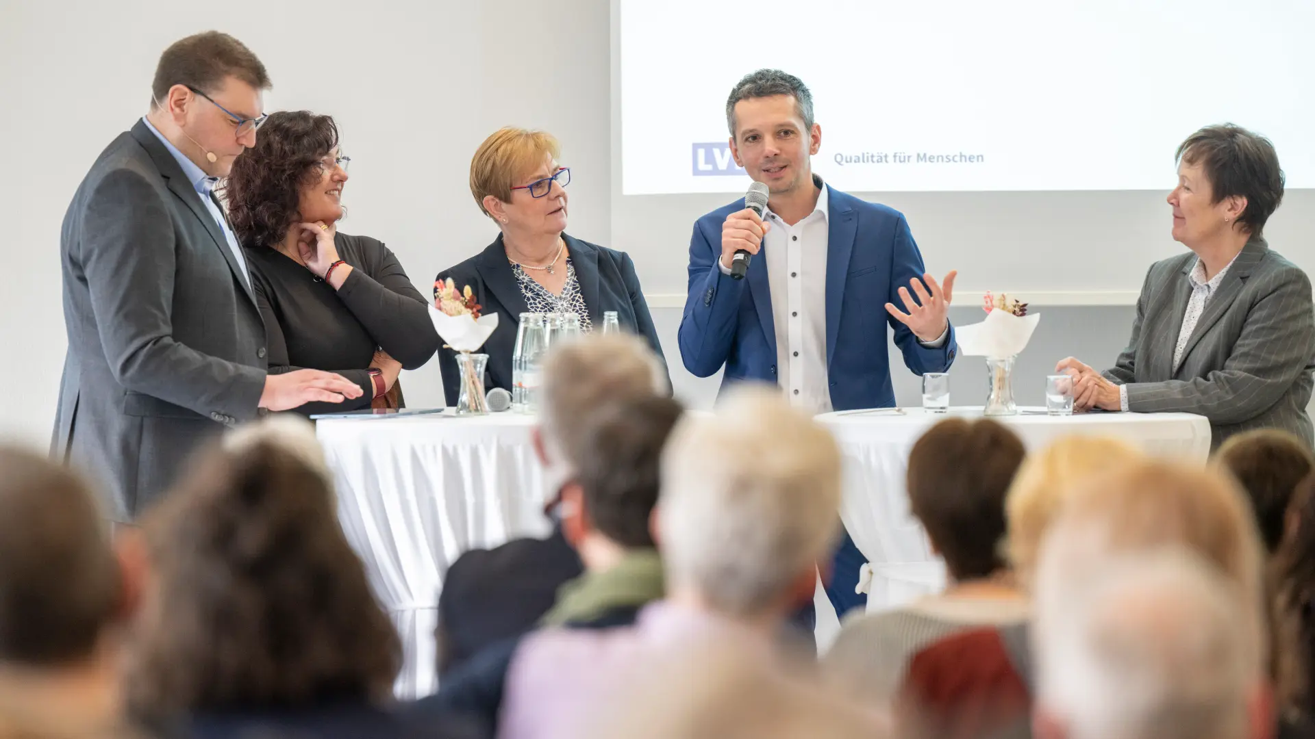 Fünf Personen stehen an einem Stehtisch, ein Mann spricht mit Mikrofon, Publikum im Vordergrund unscharf.