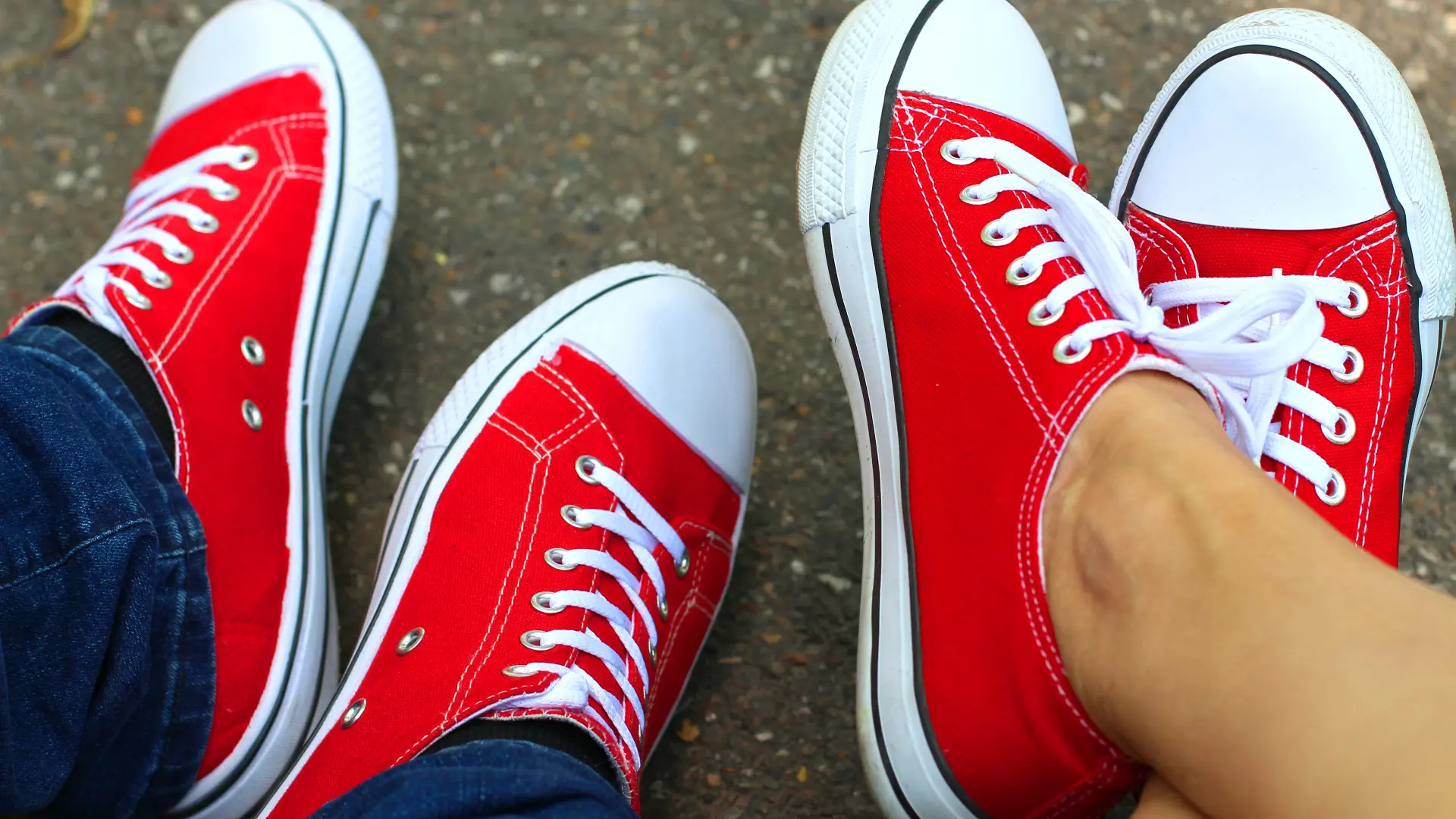 Zwei Personen tragen rote Sneakers mit weißen Sohlen und weißen Schnürsenkeln, eine Person trägt blaue Jeans, die andere hat nackte Beine.