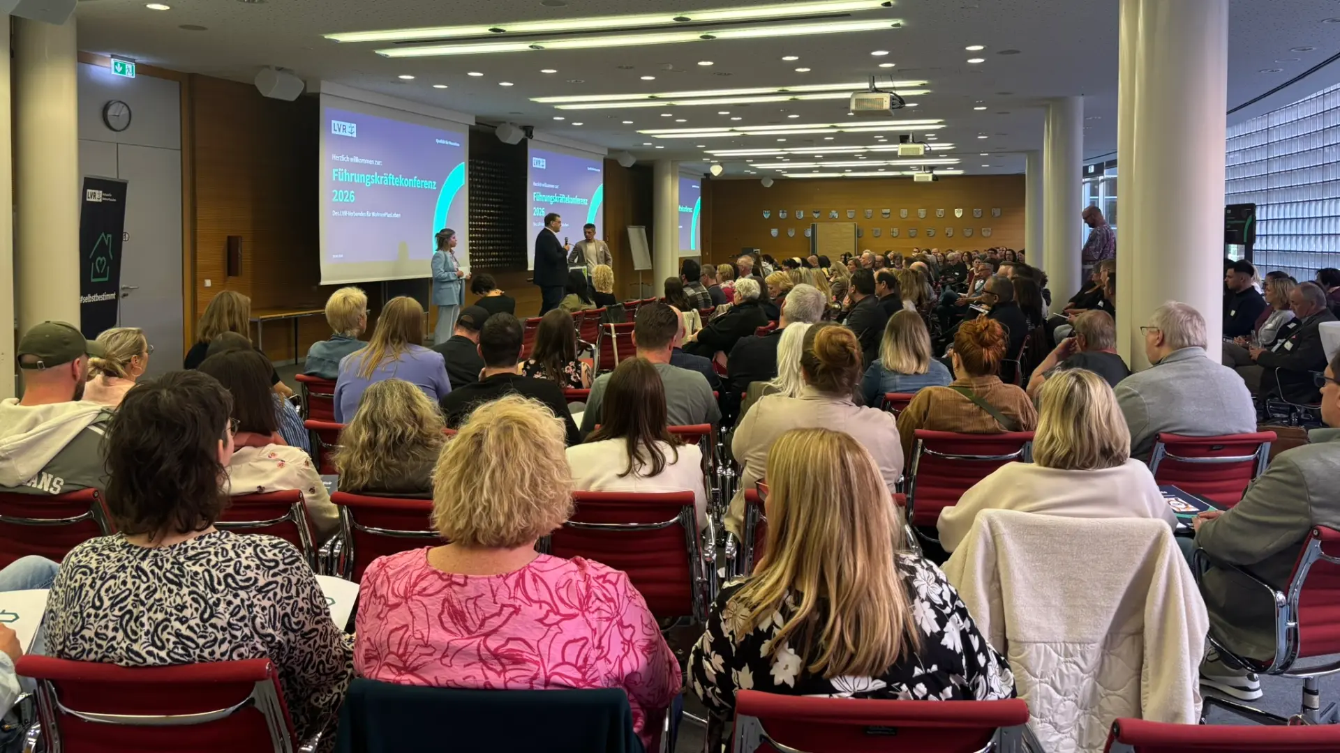 Großer Saal mit Publikum, das auf eine Bühne mit drei Personen und zwei Projektionsflächen blickt.