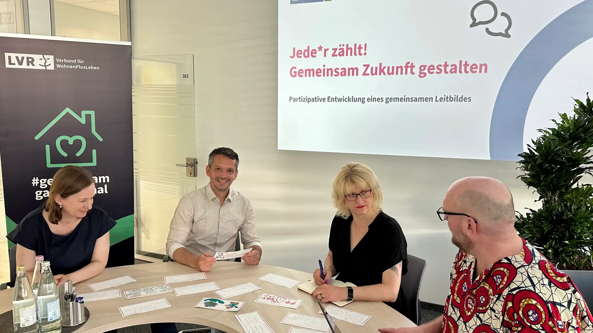 Vier Personen sitzen an einem runden Tisch in einem Besprechungsraum, im Hintergrund ein Bildschirm mit der Präsentation 'Jede*r zählt! Gemeinsam Zukunft gestalten' und ein Banner mit dem Logo von LVR