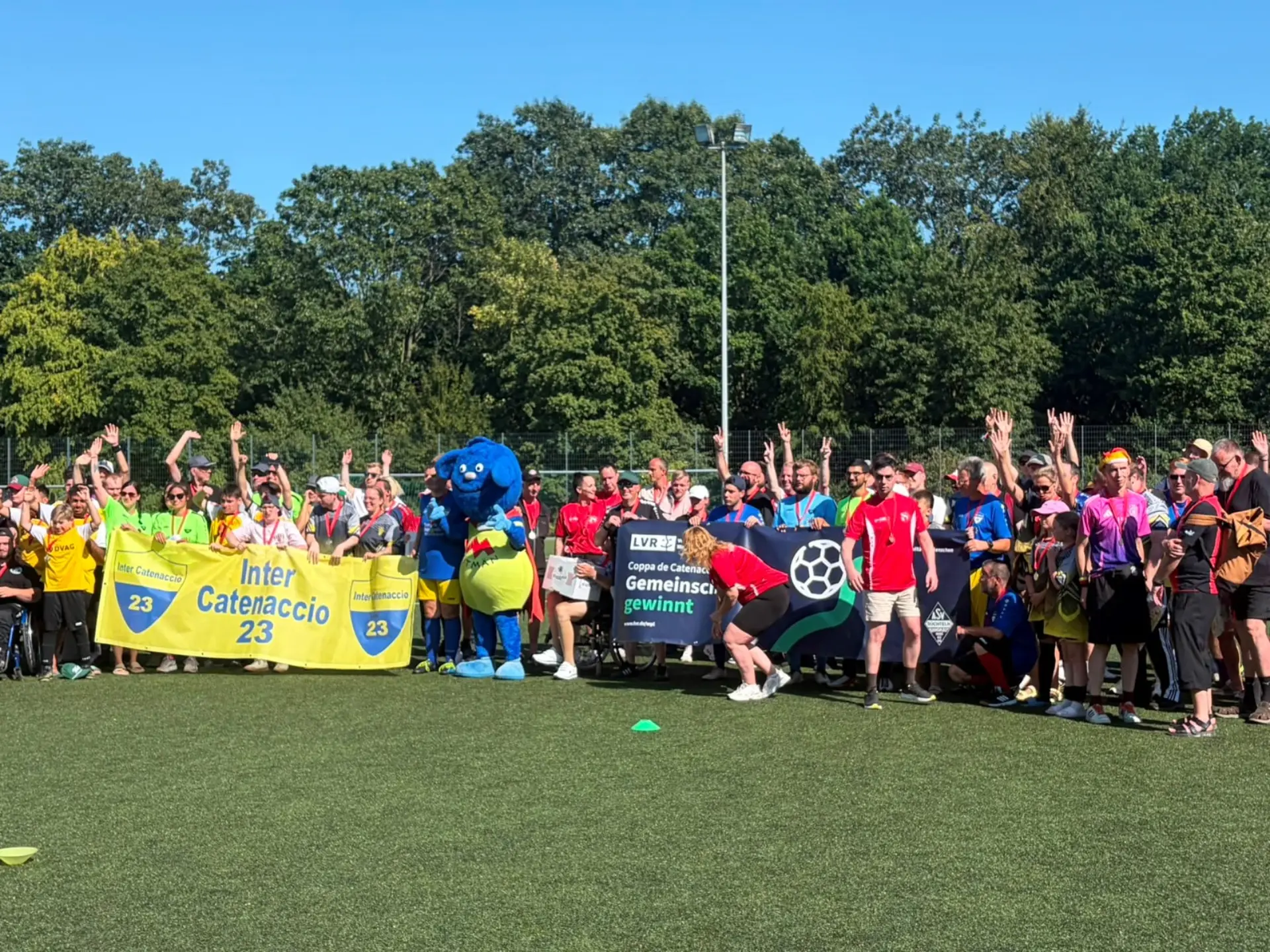 Gruppenfoto von Menschen auf einem Sportfeld mit zwei großen Bannern und einer blauen Maskottchenfigur