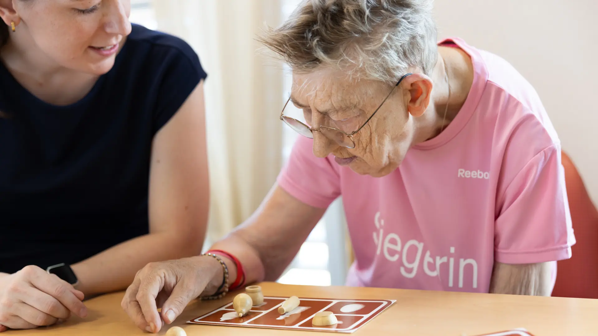 Ältere Frau mit Brille und rosa T-Shirt spielt mit Holzfiguren an einem Tisch, neben ihr sitzt eine jüngere Frau in schwarzem T-Shirt