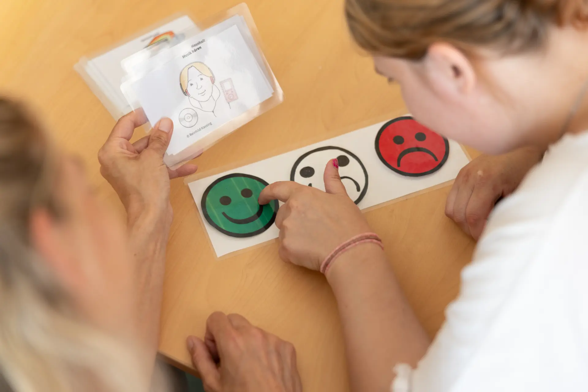 Zwei Personen sitzen an einem Tisch, eine zeigt mit dem Finger auf ein grünes Smiley-Symbol auf einer Reihe von drei Smiley-Karten mit unterschiedlichen Gesichtsausdrücken.