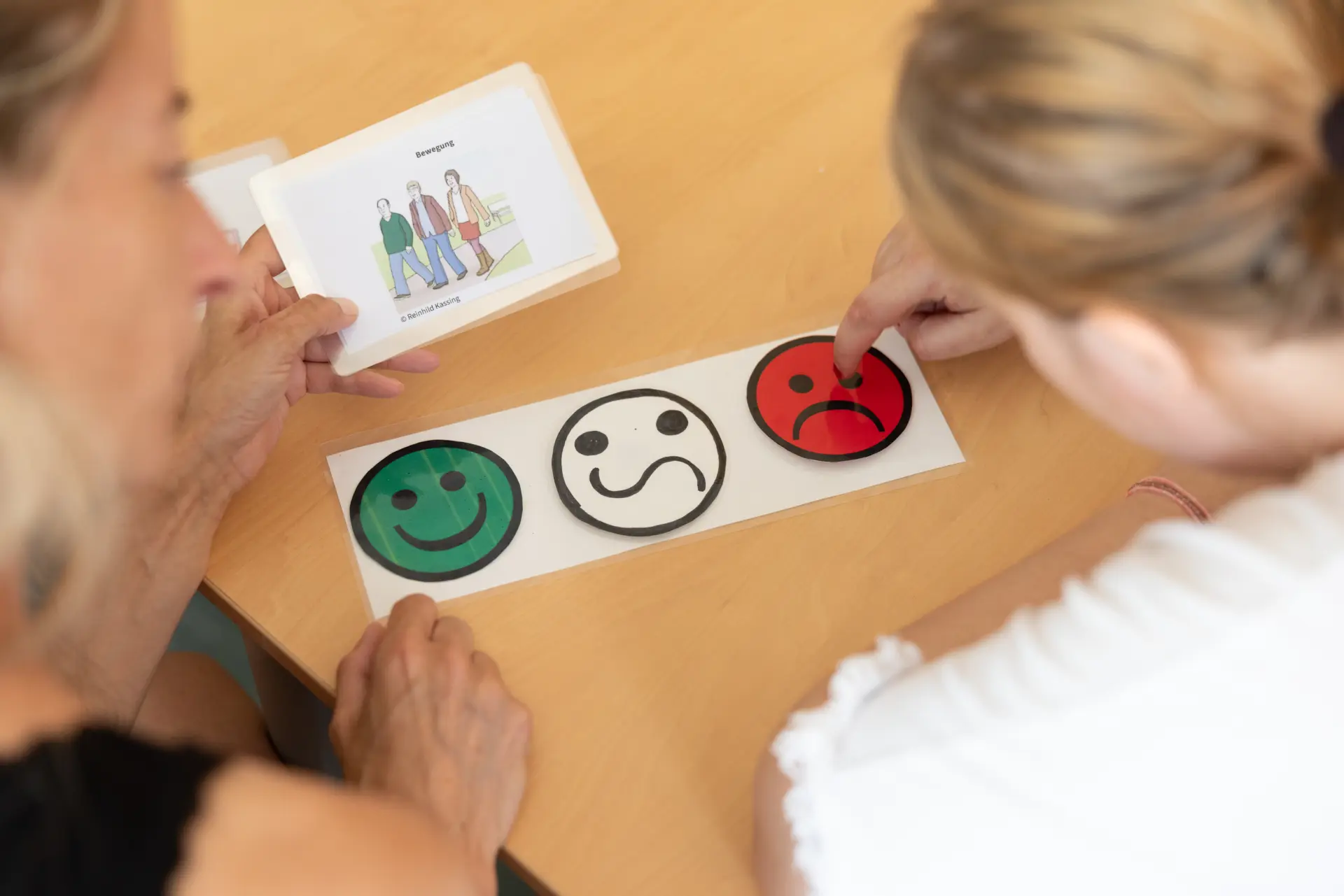 Zwei Personen sitzen an einem Tisch, eine hält eine Karte mit einer Illustration, die andere zeigt auf ein rotes, trauriges Smiley auf einer Reihe von drei Smileys in grün, weiß und rot