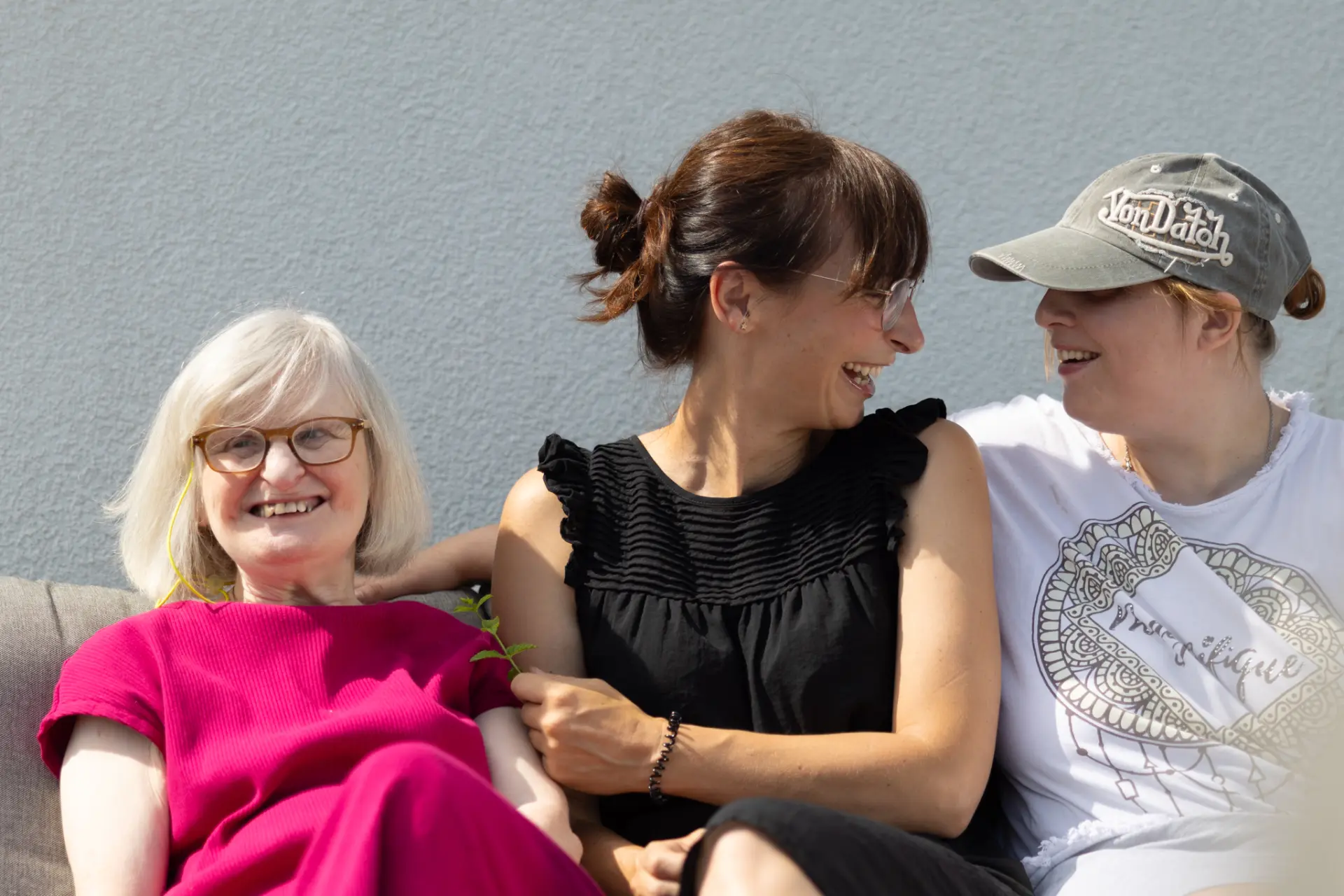 Drei Frauen sitzen eng nebeneinander auf einer Couch, die linke trägt ein pinkes Kleid, die mittlere ein schwarzes Kleid, die rechte ein weißes T-Shirt und eine graue Kappe.