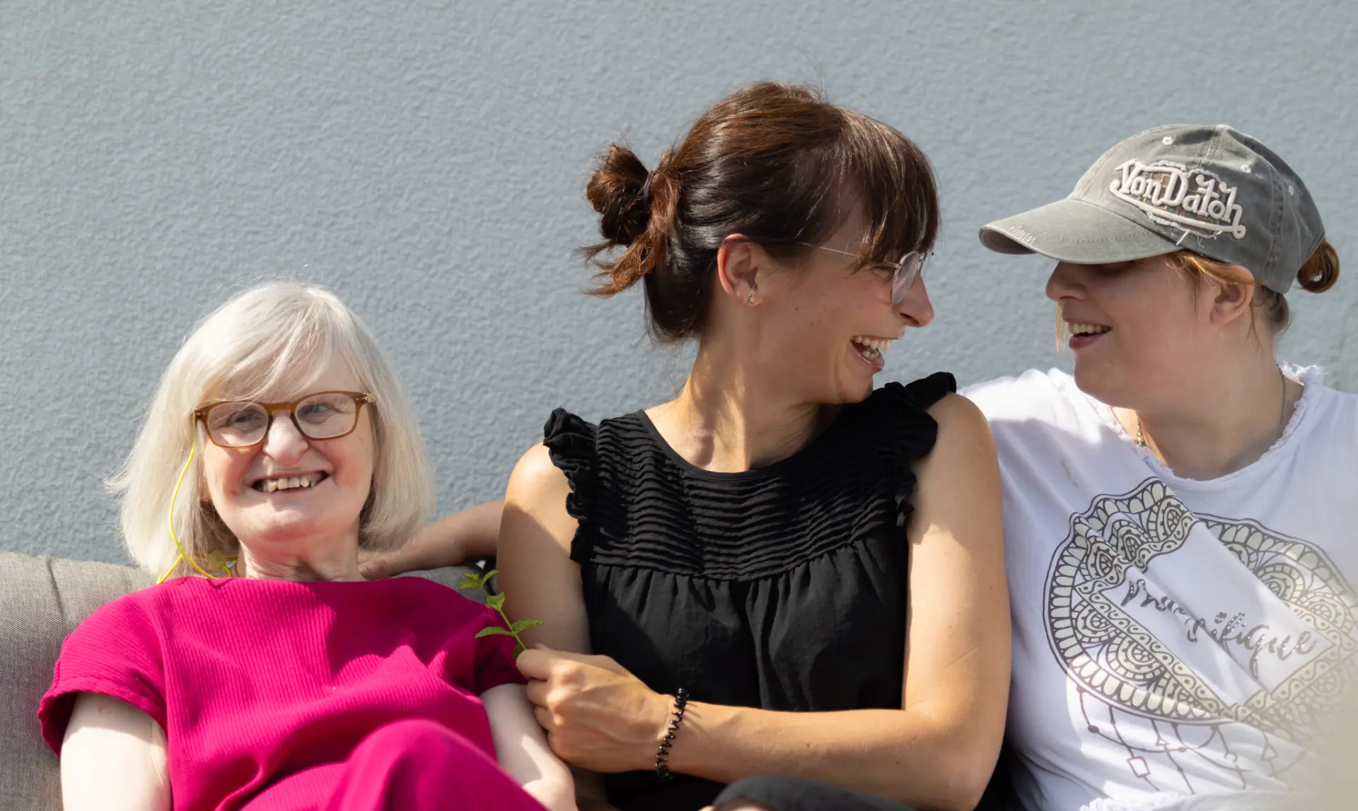 Drei Frauen sitzen eng nebeneinander auf einer Couch, die linke trägt ein pinkes Kleid, die mittlere ein schwarzes Kleid, die rechte ein weißes T-Shirt und eine graue Kappe.