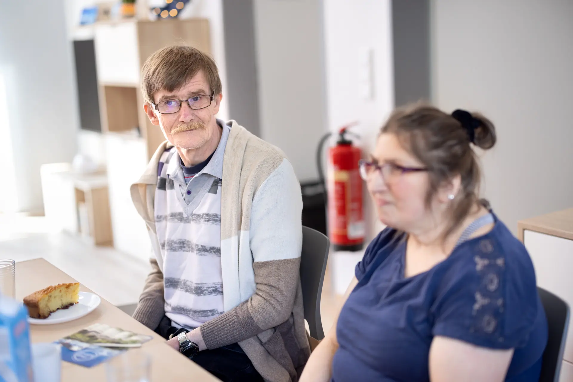 Zwei sitzende Personen an einem Tisch, ein Mann mit Brille und gestreiftem Pullover blickt zur Seite, eine Frau mit Brille und blauem Oberteil sitzt daneben