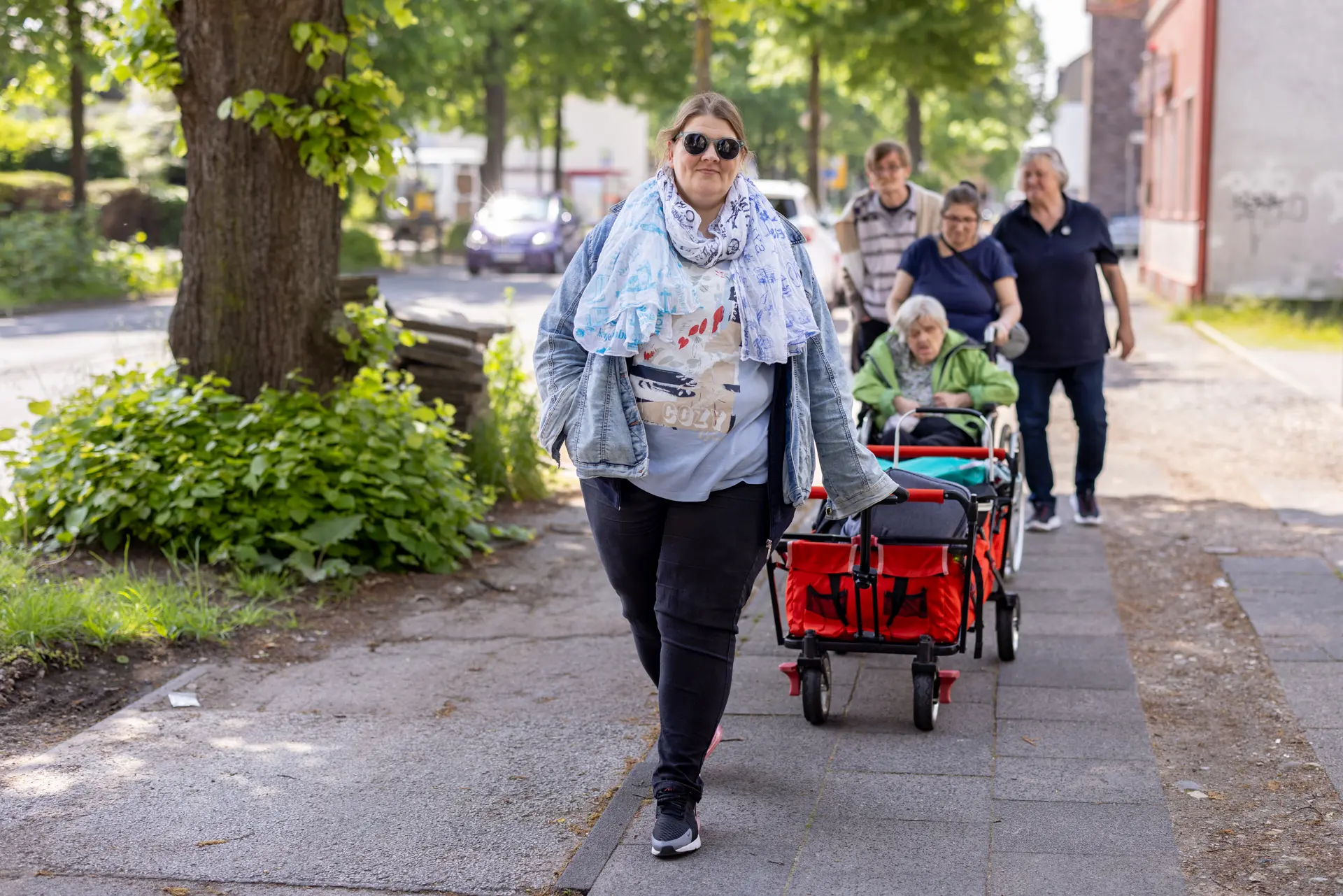 Frau mit Sonnenbrille und Schal zieht roten Bollerwagen auf einem Gehweg, dahinter folgen vier weitere Personen, eine davon sitzt im Bollerwagen