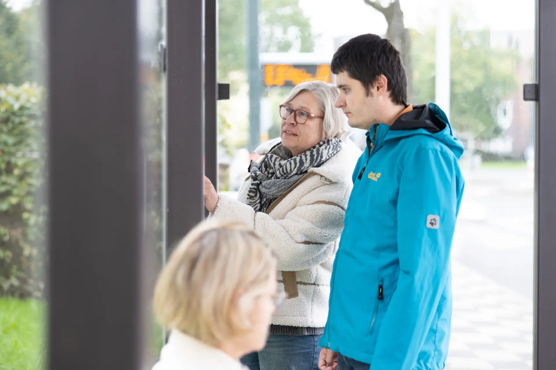 Ältere Frau mit Brille und Schal zeigt einem jungen Mann in blauer Jacke etwas auf einer Informationstafel an einer Haltestelle