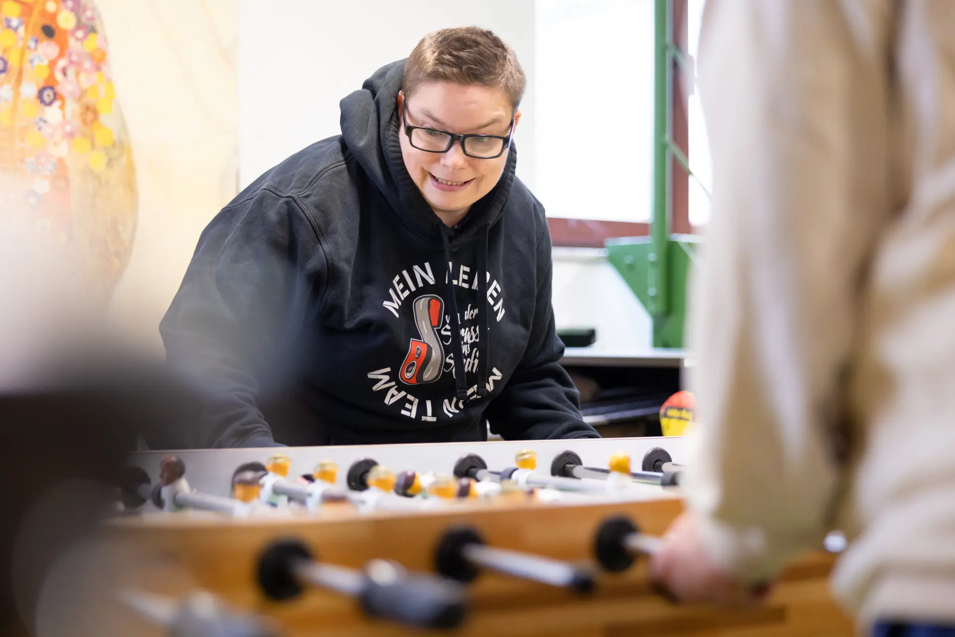 Person mit Brille und Kapuzenpullover spielt Tischfußball in einem hellen Raum
