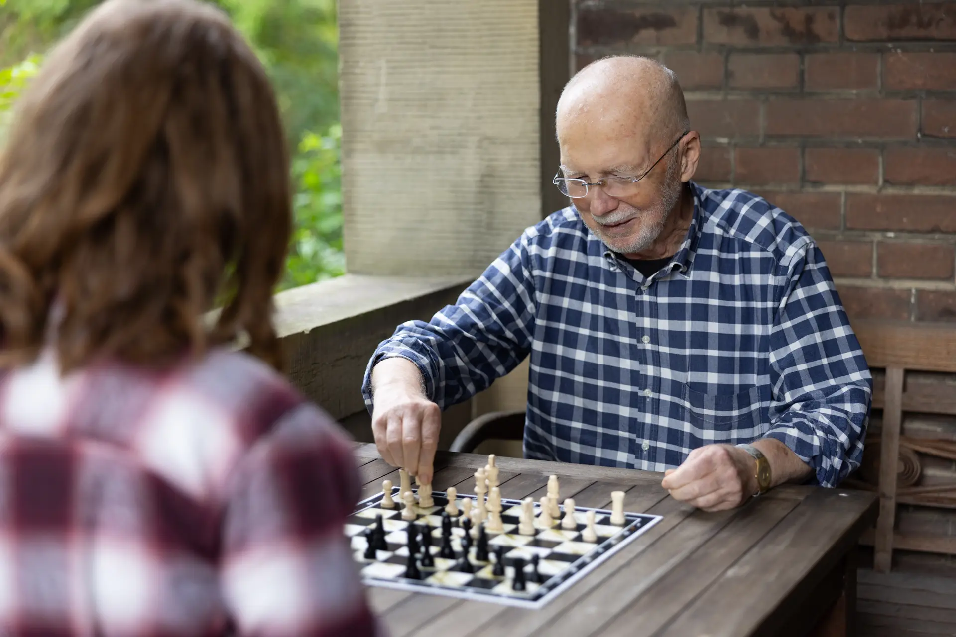 Älterer Mann mit Brille und kariertem Hemd spielt Schach mit einer jüngeren Person an einem Holztisch im Freien