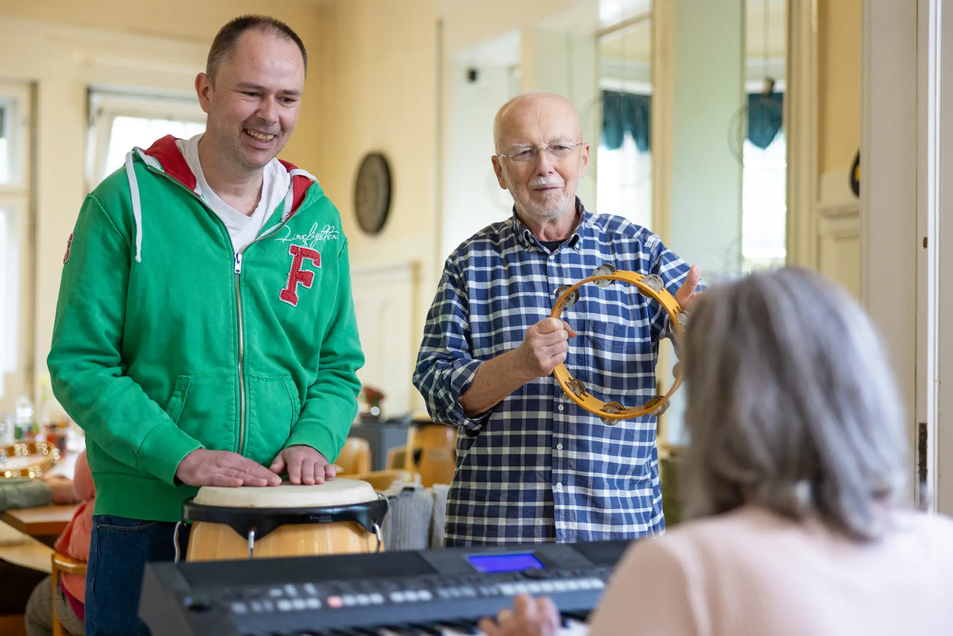 Zwei Männer musizieren gemeinsam, einer spielt Bongos, der andere hält ein Tamburin, im Vordergrund ist eine Person an einem Keyboard zu sehen