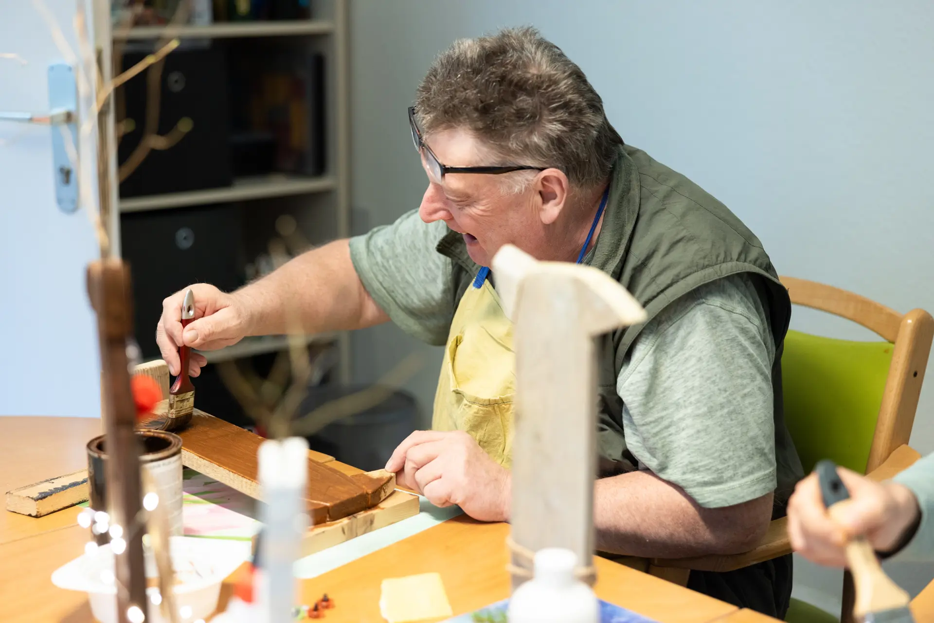 Mann mit Brille und grünem T-Shirt sitzt an einem Tisch und arbeitet mit einem Pinsel an einem Holzstück