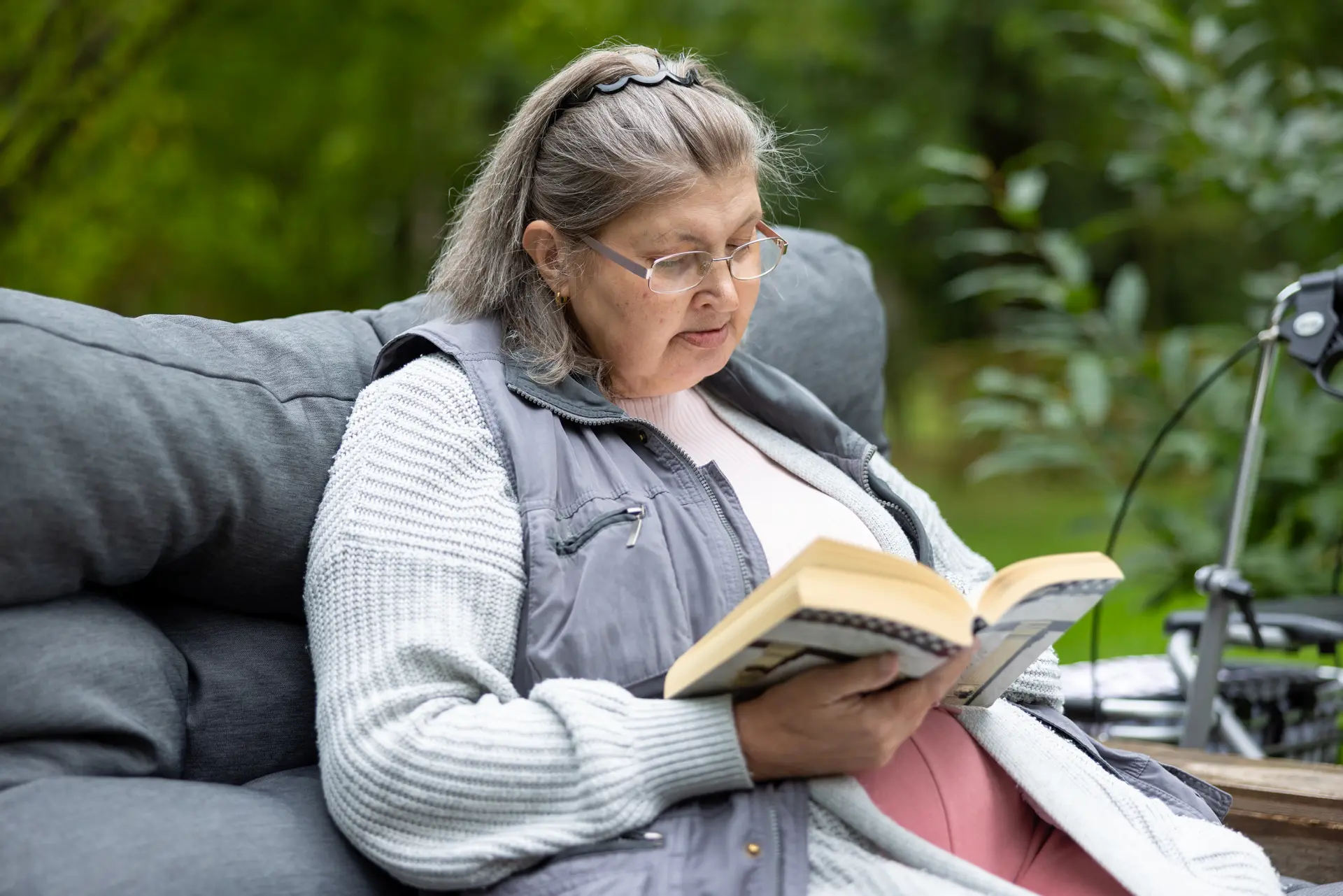 Ältere Frau mit Brille und grauen Haaren liest auf einer Couch im Freien ein Buch
