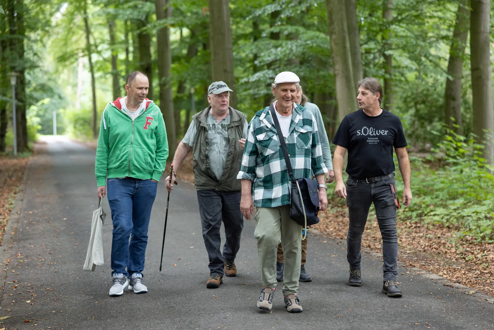 Fünf Männer gehen auf einem Waldweg, einer trägt einen Gehstock, ein anderer eine weiße Tasche, sie sind locker gekleidet und unterhalten sich.