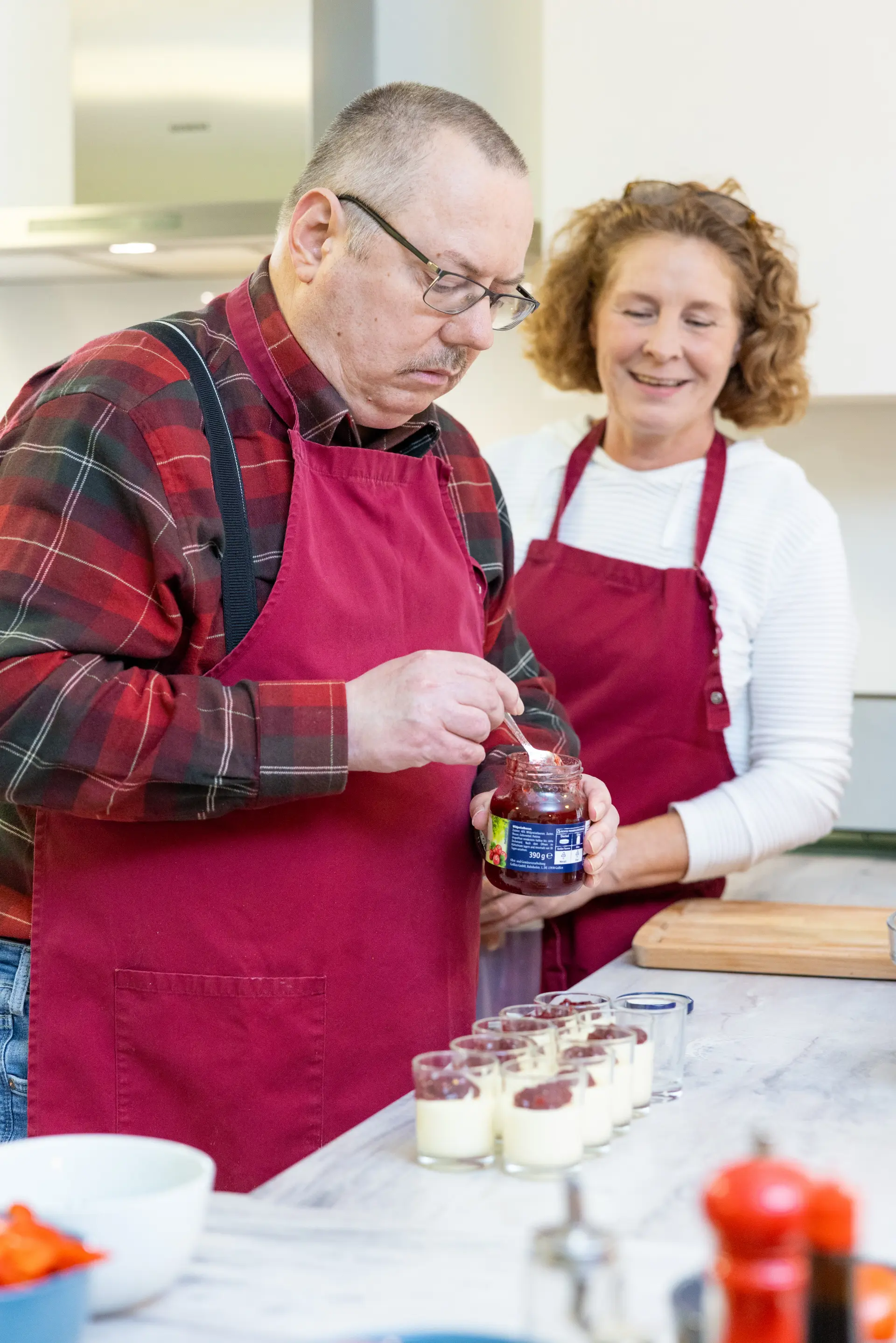 Zwei Personen mit roten Schürzen in einer Küche, eine hält ein Glas mit roter Marmelade, die andere steht daneben