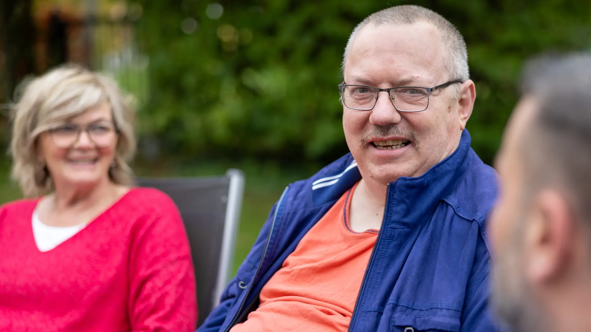 Drei Personen sitzen im Freien, zwei von ihnen sind unscharf im Hintergrund, eine trägt ein rotes Oberteil, eine andere ein orangefarbenes Shirt mit blauer Jacke.