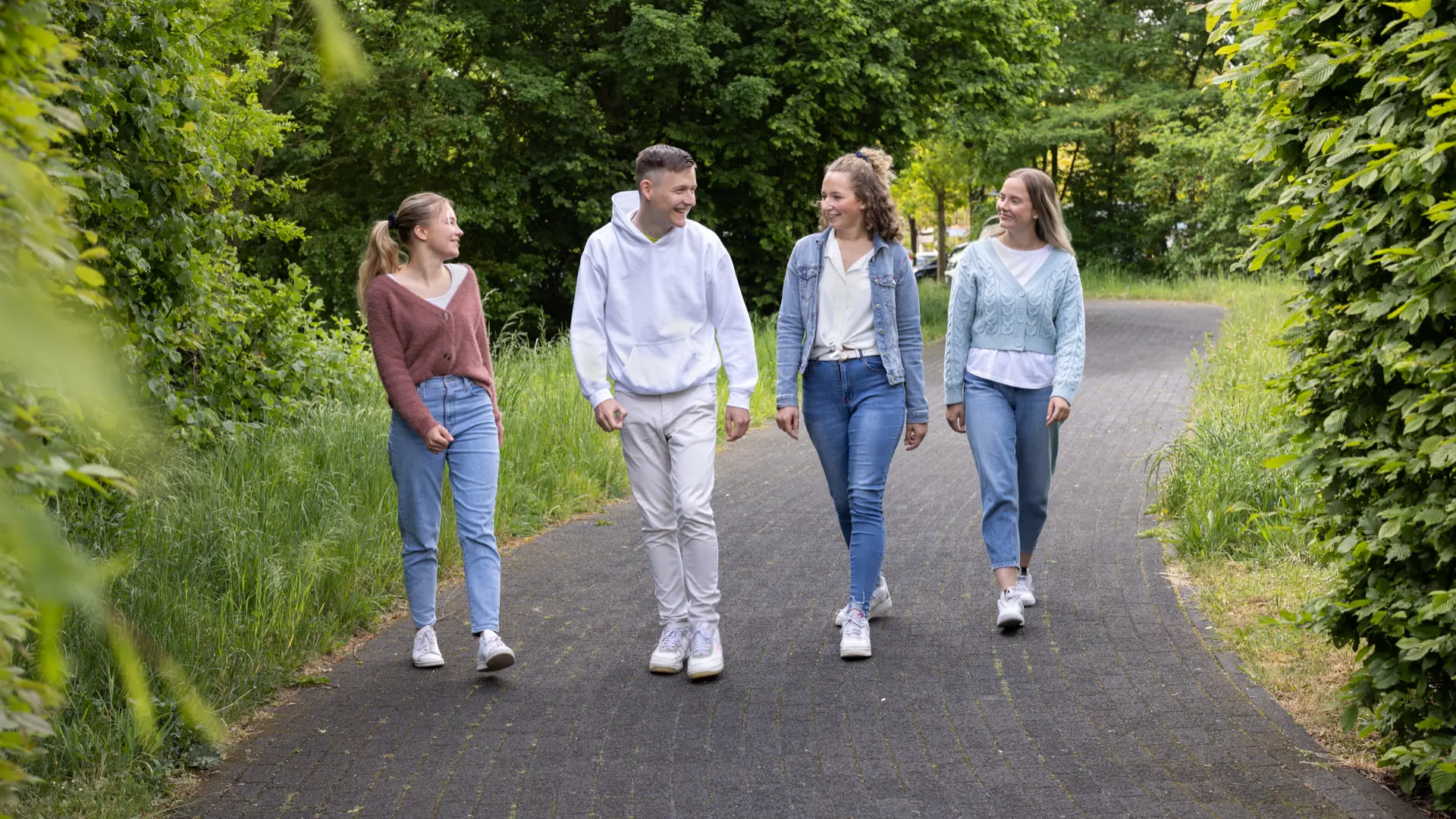 Vier junge Menschen gehen auf einem gepflasterten Weg in einem grünen Park und unterhalten sich dabei