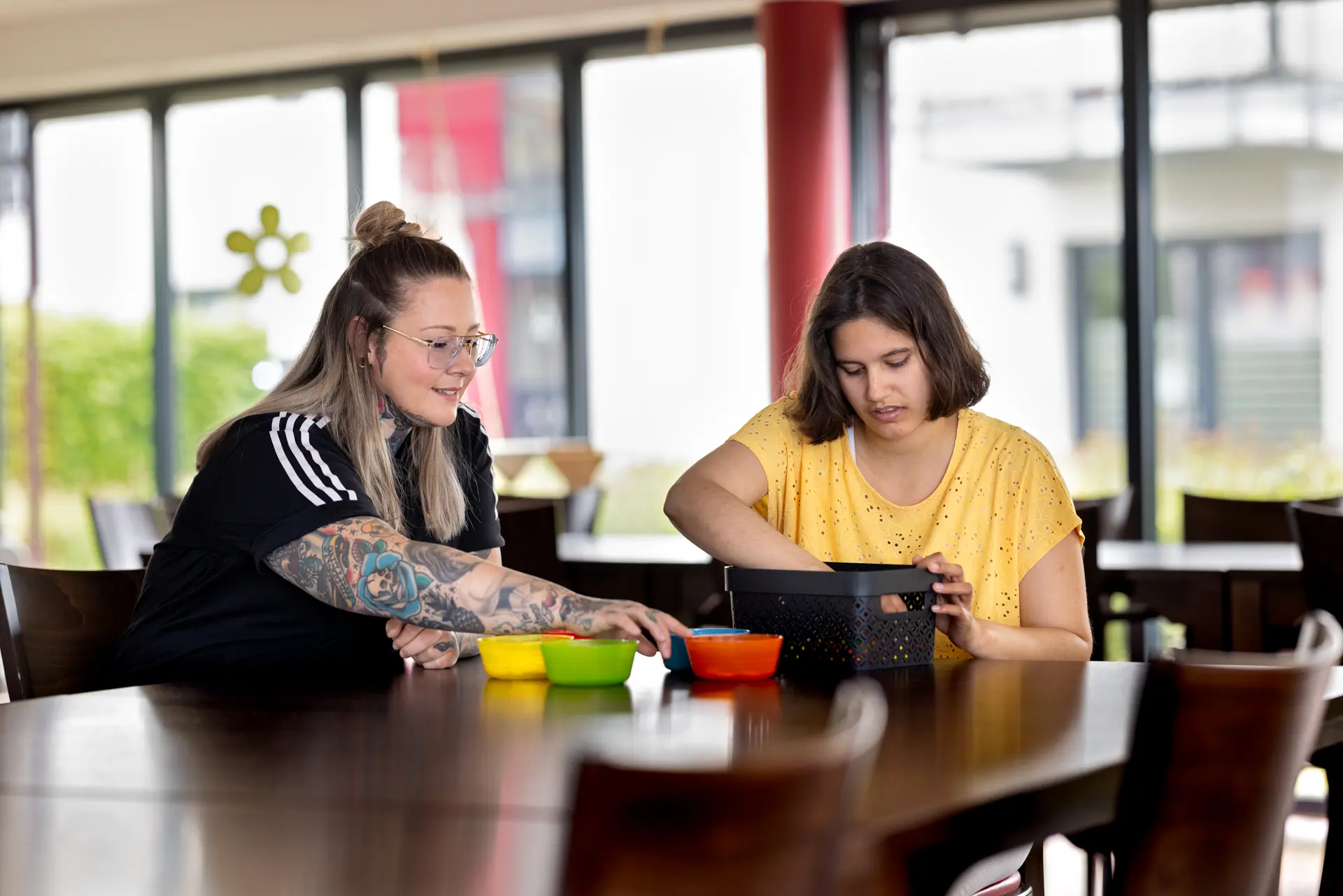 Zwei Frauen sitzen an einem Tisch, eine mit Brille und Tattoos zeigt auf bunte Schalen, die andere öffnet eine schwarze Box