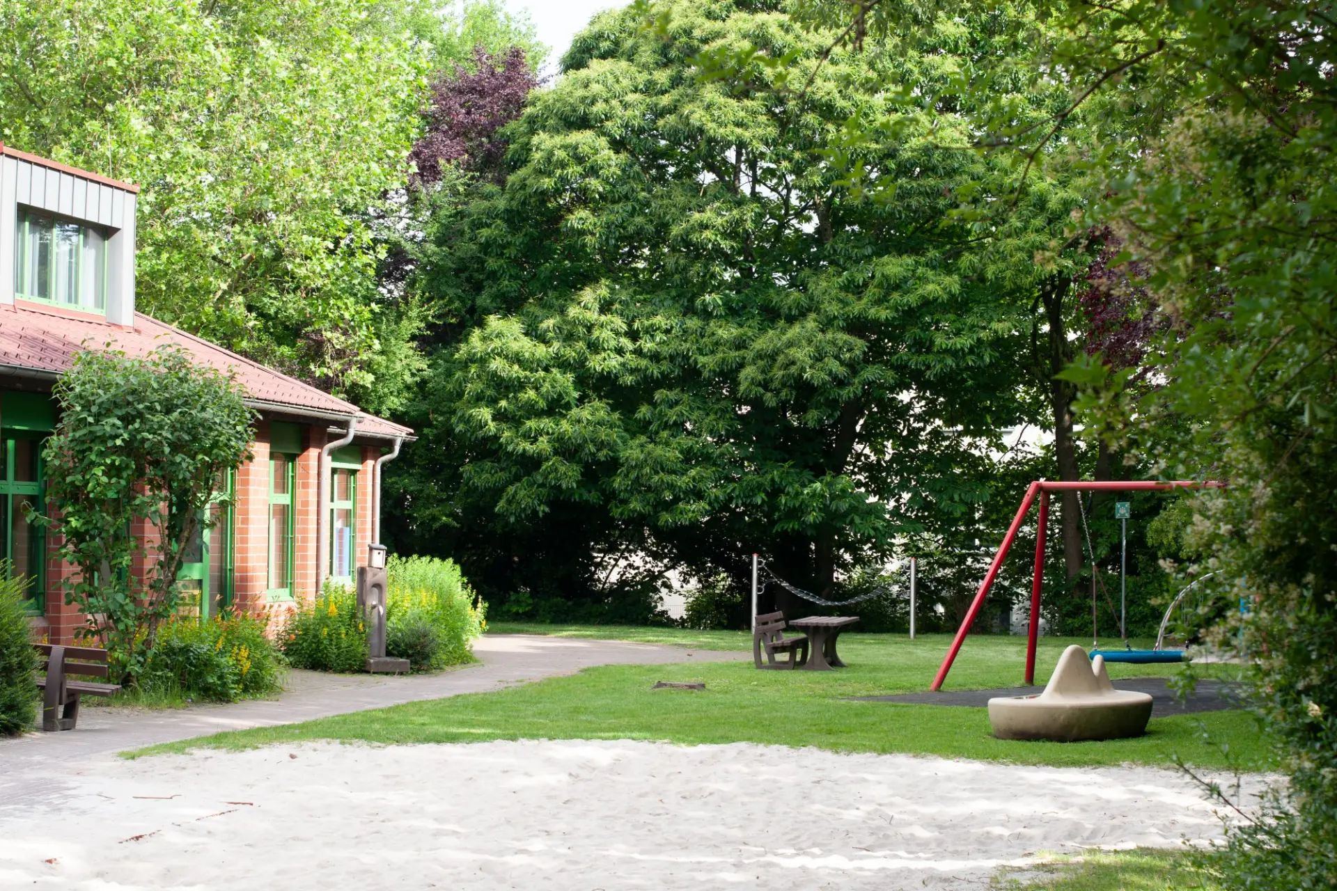 Foto vom sehr grünen Innenhof mit einem Schulgebäude links, Tisch, Bänke und Schaukel rechts