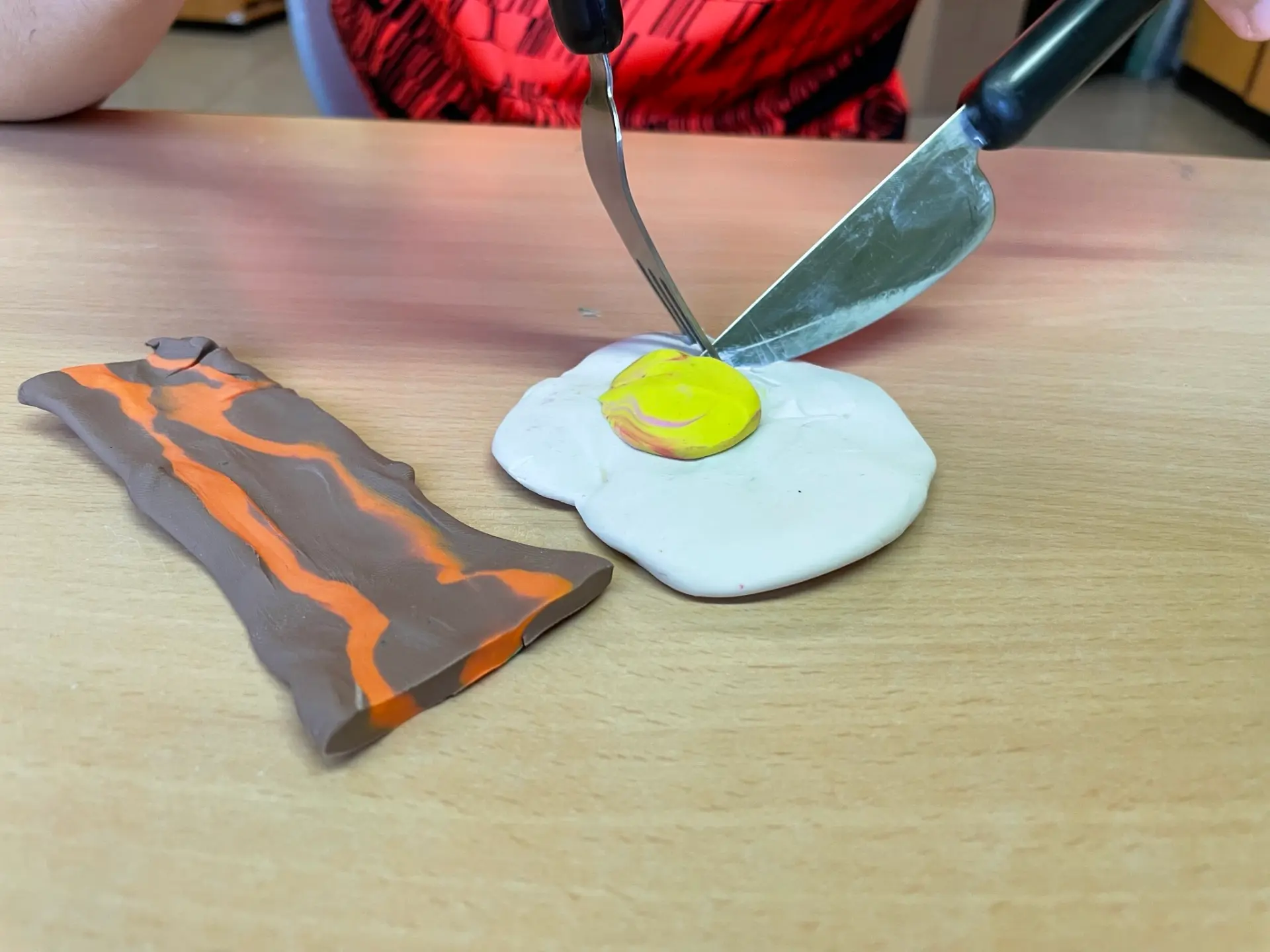 Die Hände von einem Kind, das an einem Spiegelei und Baconstück aus Knete das Essen mit Messer und Gabel übt.