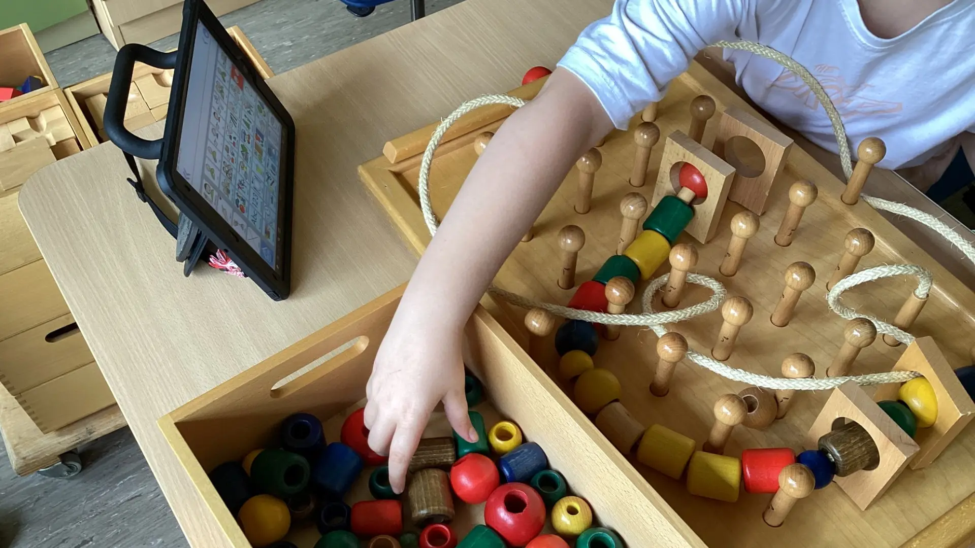 Ein Kind spielt mit einem motoirischen Holzteilespiel am Tisch, an der Seite ist ein Tablet mit Metacom-Symbolen.