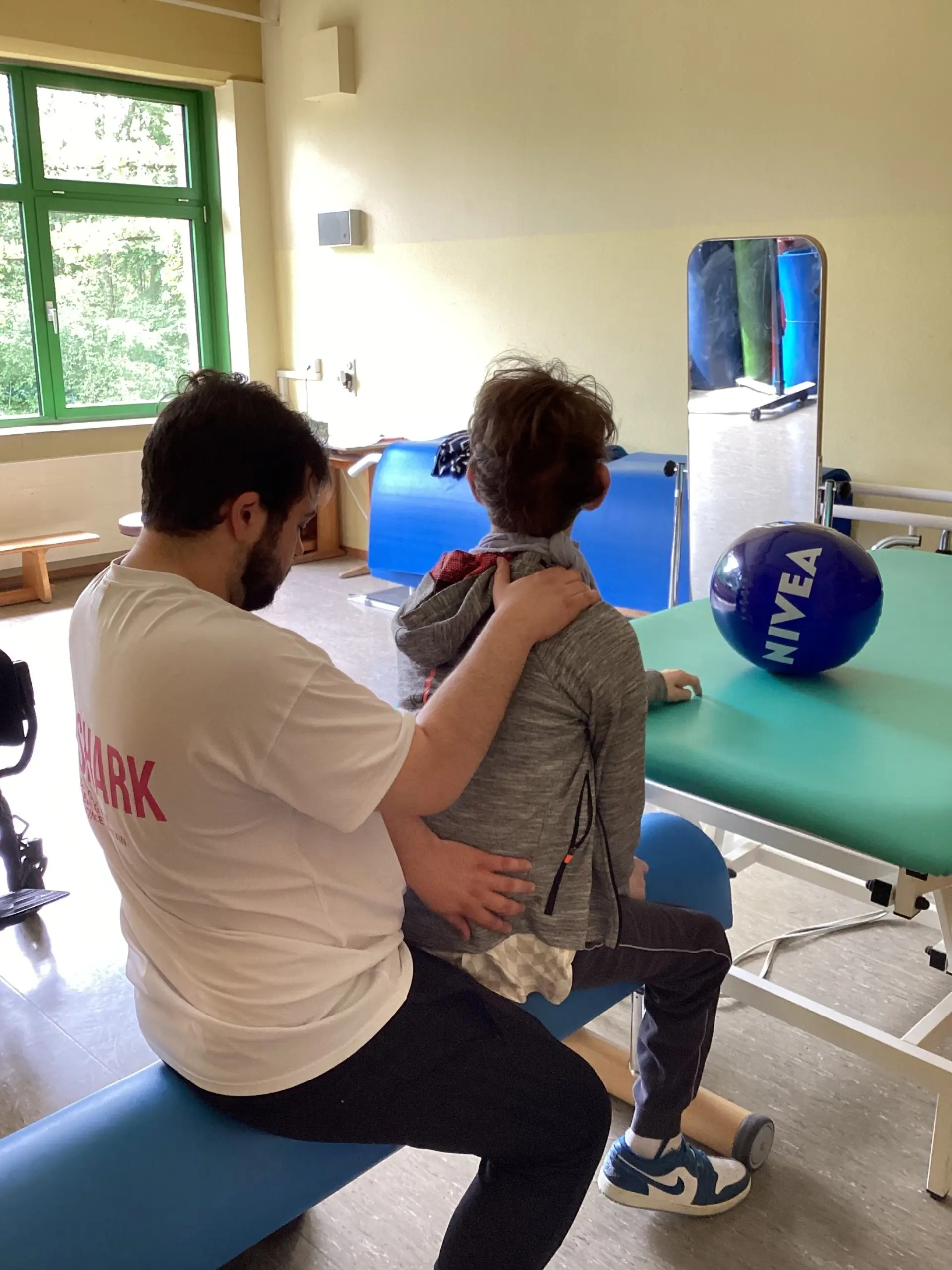 Im Physiotherapieraum der Schule: Ein Physiotherapeut behandelt den Rücken eines Schülers vor ihm.