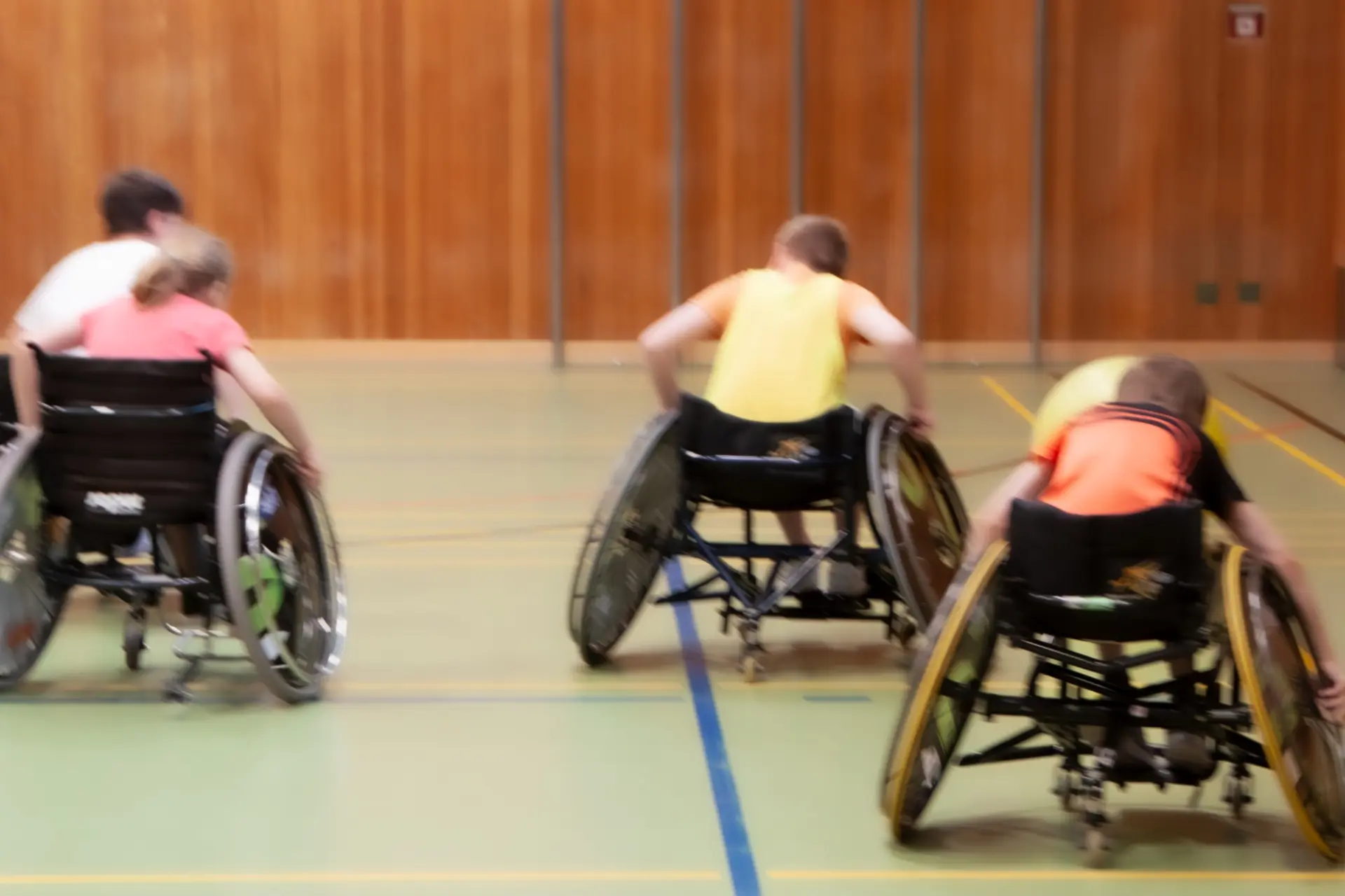 Vier Kinder im Rollstuhl fahren schnell in Richtung eines großen Balls in der Turnhalle.