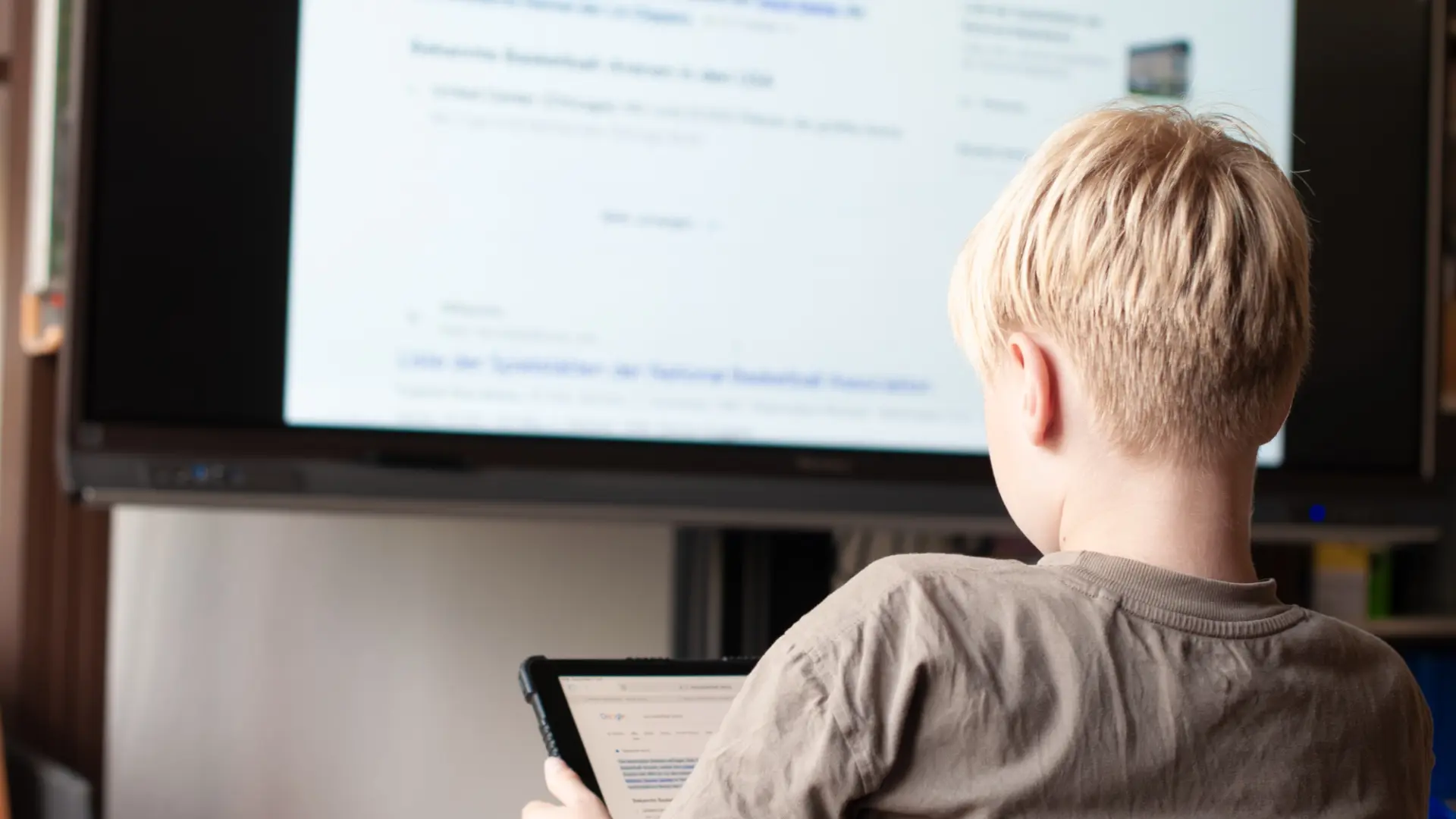 Ein Schüler schaut auf seinem Tablet und liest. An der digitalen Tafel vor ihm wird der gleiche Text angezeigt.