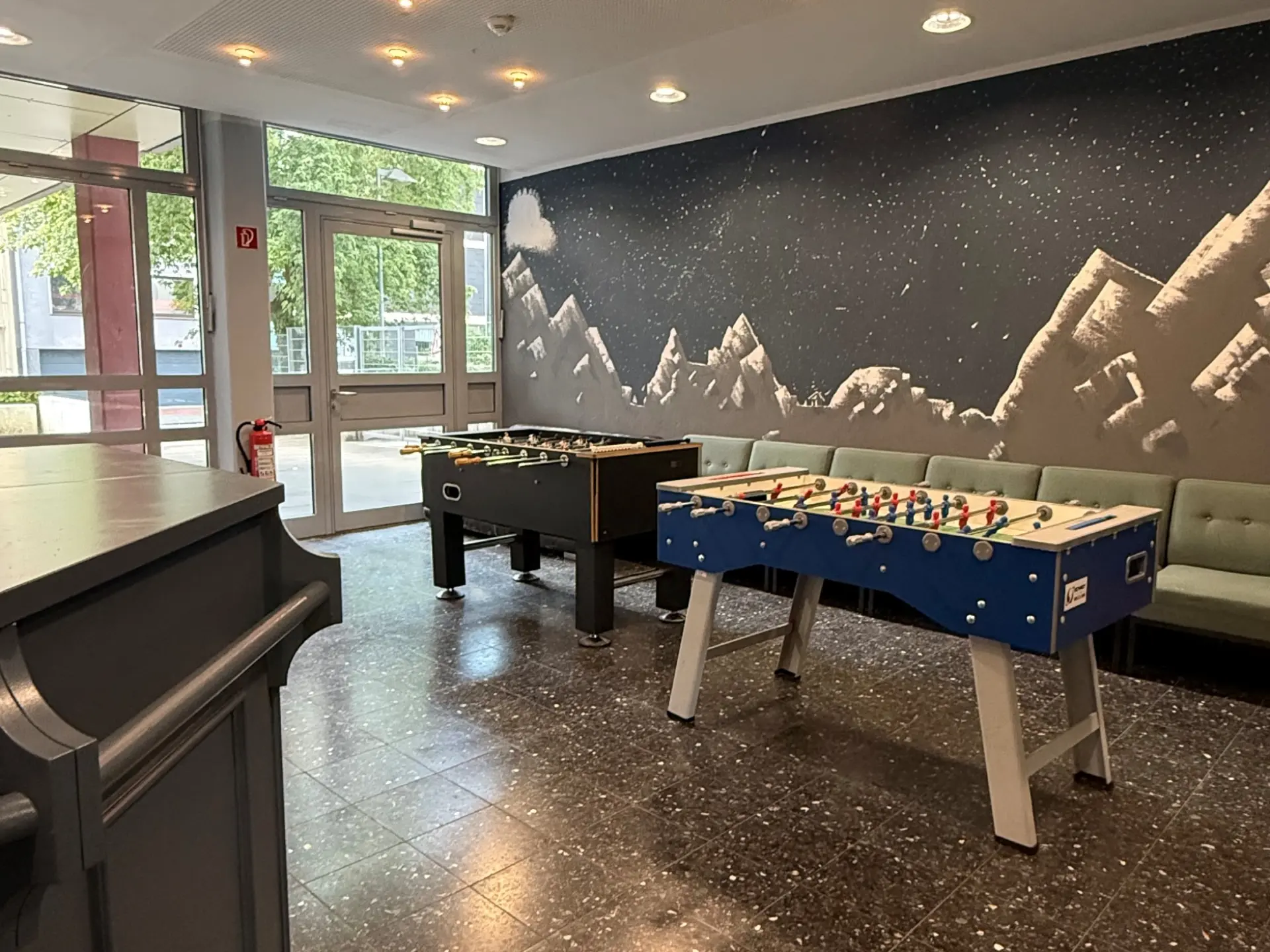Ein gemütlicher Freizeitraum mit großer Fensterfront, zwei Kickertischen, einer Sitzbank und einer großen Wandbemalung mit Bergmotiv.
