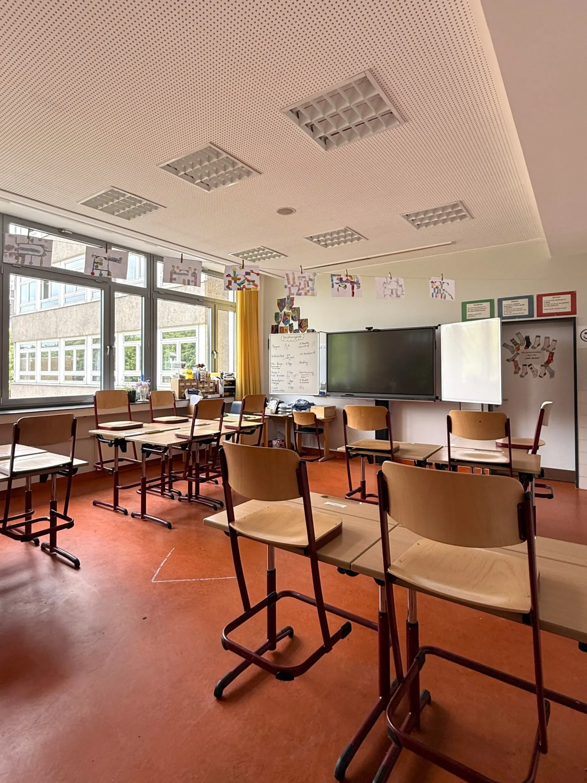 Heller Klassenraum der WKS mit Holztischen und -stühlen, großen Fenstern sowie einer digitalen Tafel und Bildern an der Wand.