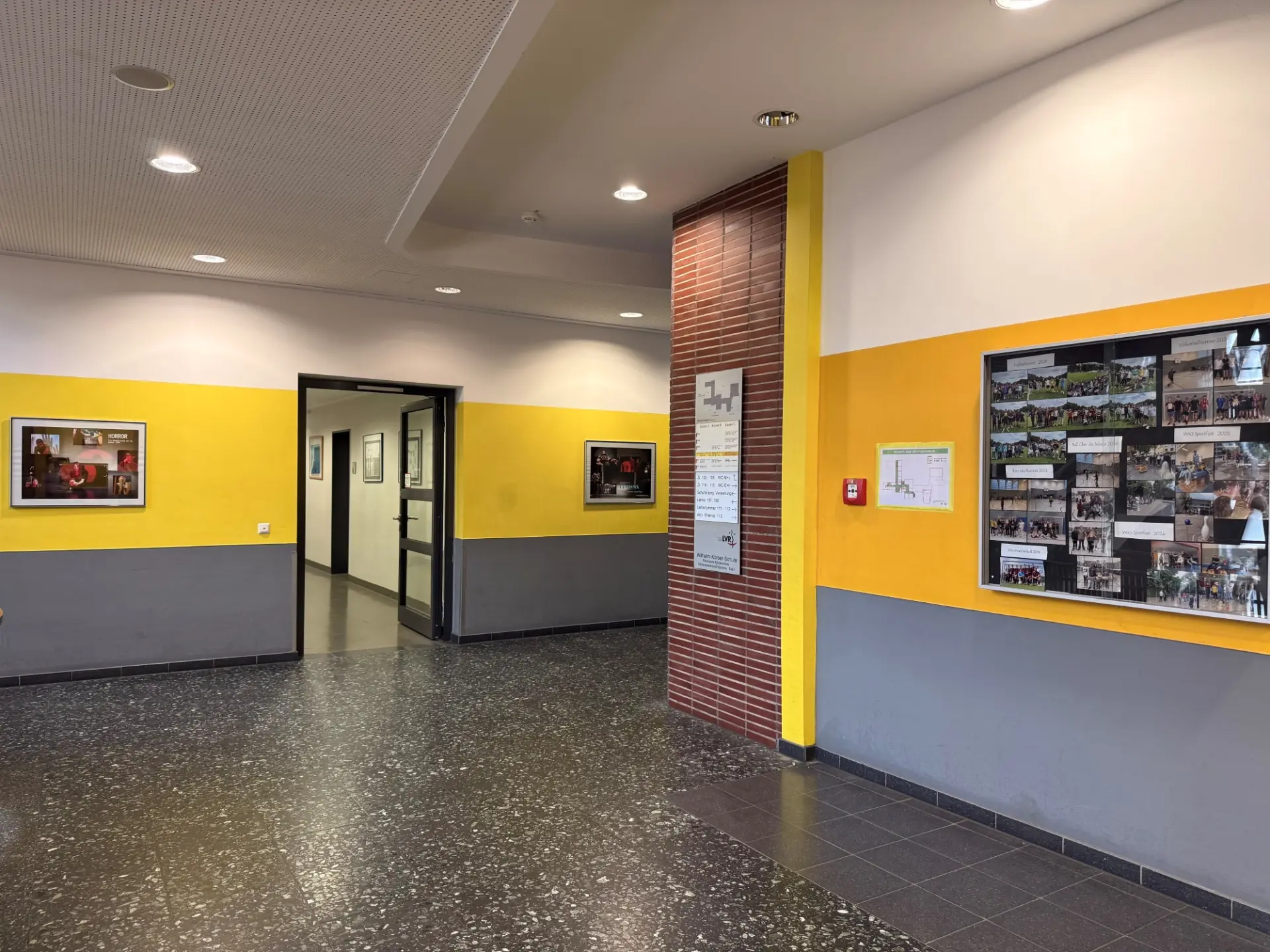 Schulflur zum Verwaltungsbereich mit gelb-grauen Wänden, einem Wegweiser und einem großen Schaukasten.