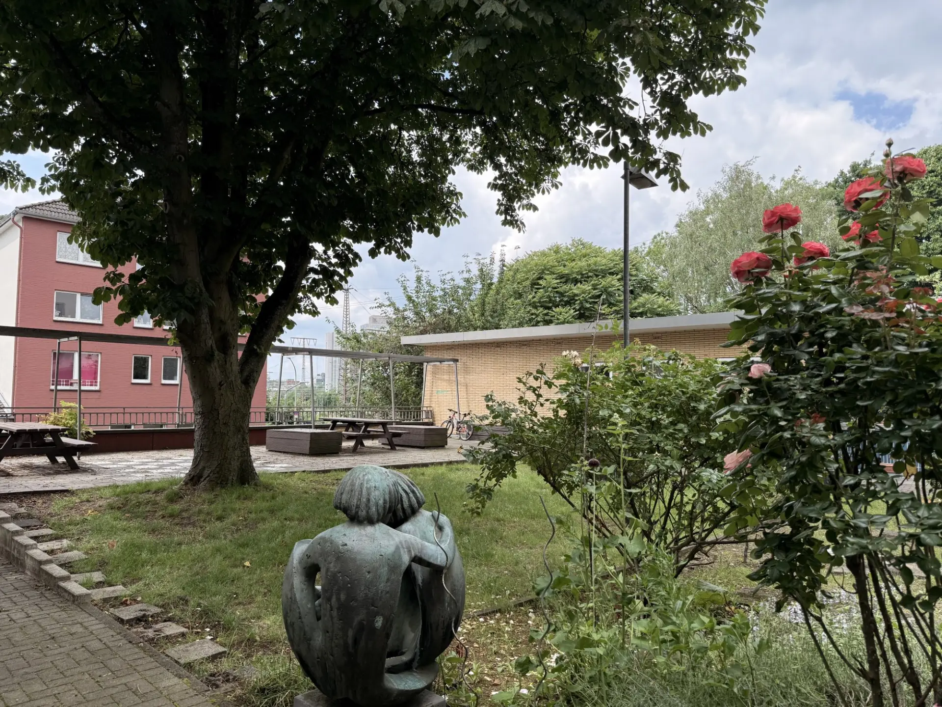 Grüner Schulgarten mit einer Skulptur, blühenden Rosen und Sitzbänken unter einem großen Baum.