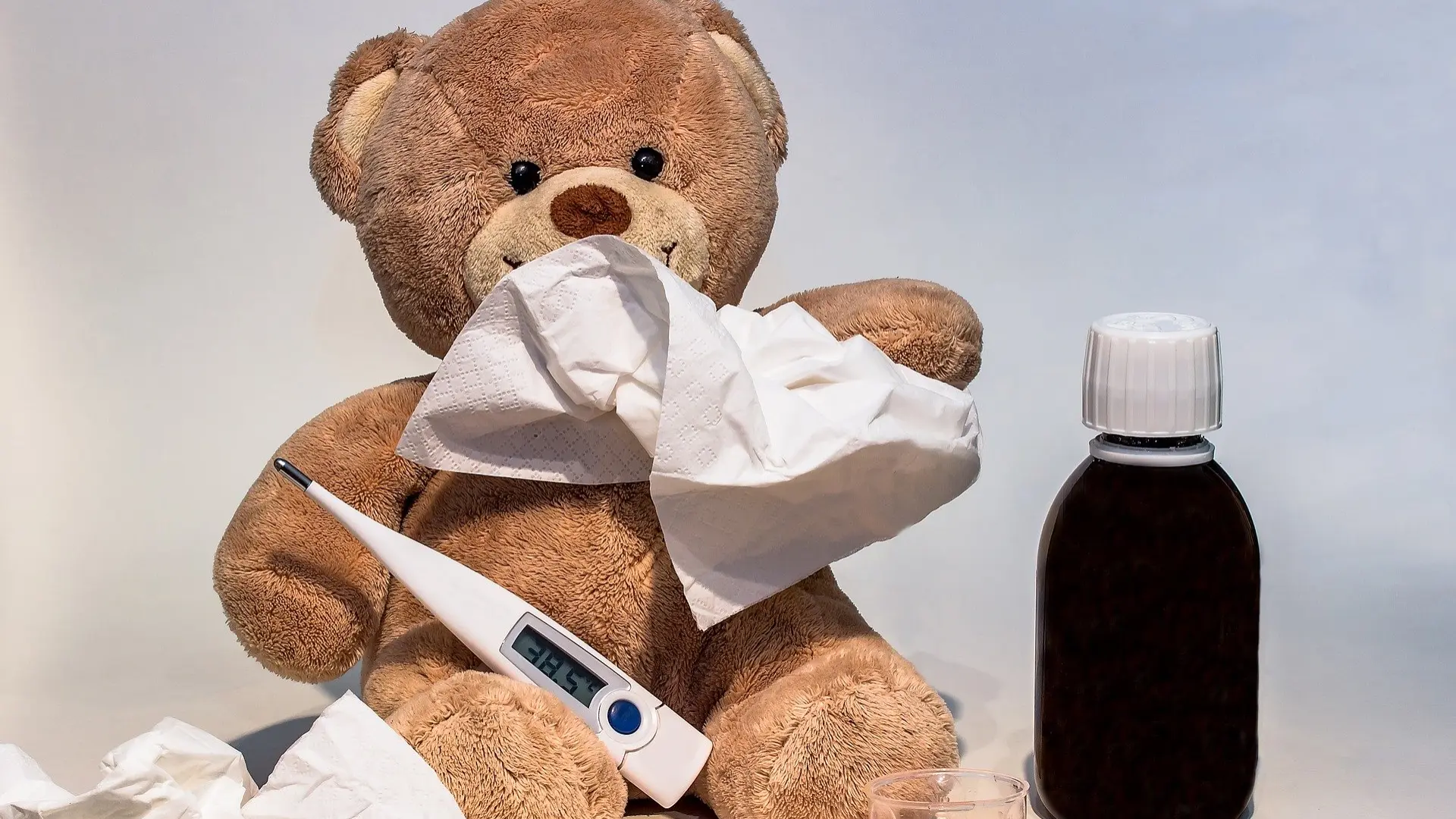 Ein Stoffbär mit Schnupfen hält sich ein Taschentuch an die Nase. Vor ihm liegen ein digitales Fieberthermometer mit der Anzeige 38,5 Grad, eine Flasche Hustensaft und ein gefüllter Messbecher.