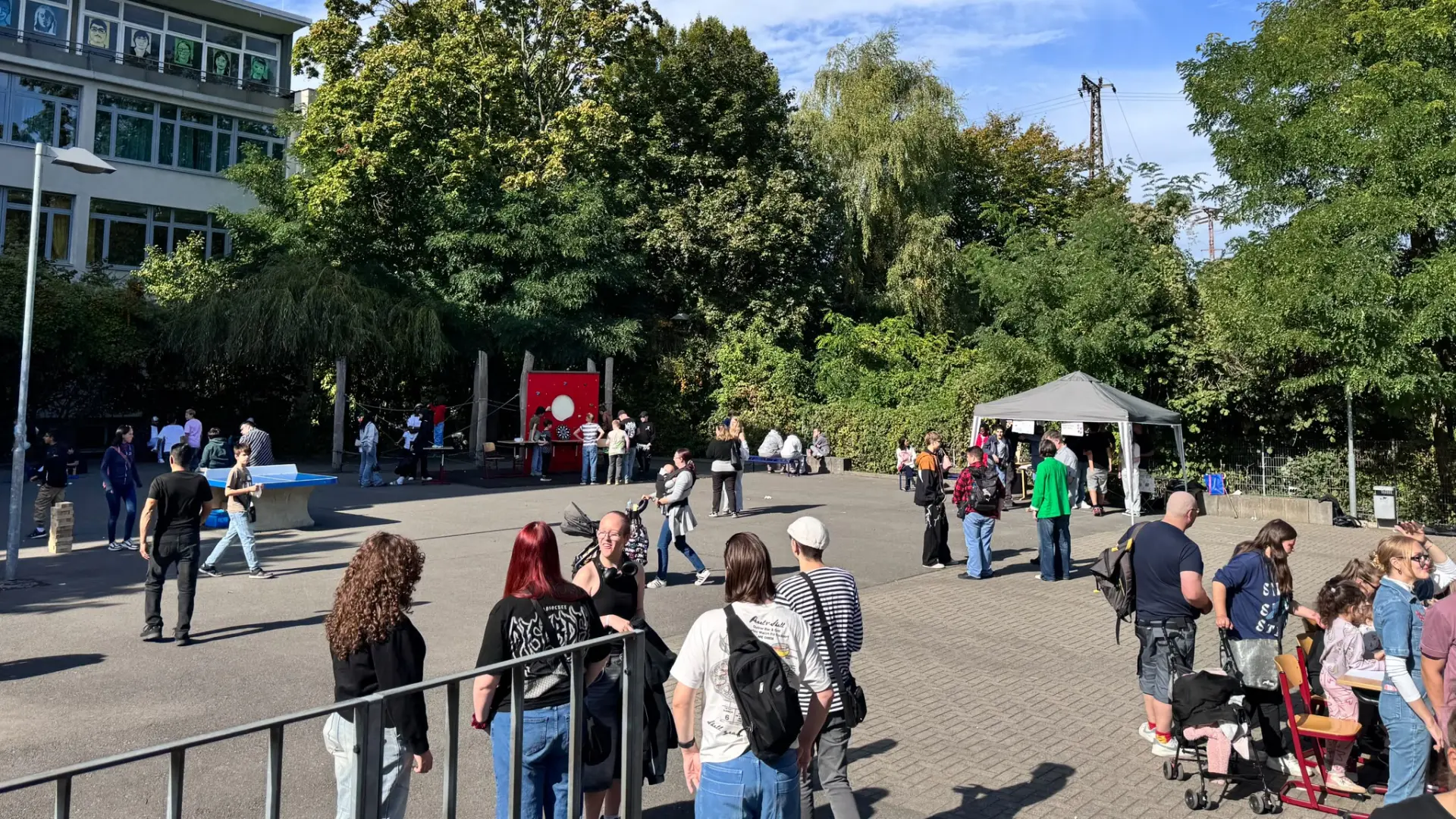 Ein weitläufiger Platz vor dem Schulgebäude der LVR-Wilhelm-Körber-Schule. Überall sind Kinder, Jugendliche und Erwachsene zu sehen. In der Mitte spielen neun Schüler*innen ein Spiel mit Seilen und Holzblöcken.