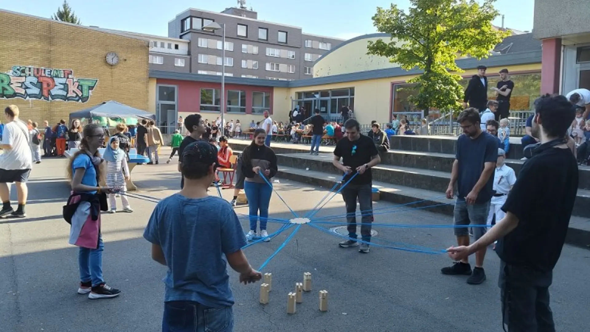 Ein weitläufiger Platz vor dem Schulgebäude der LVR-Wilhelm-Körber-Schule. Überall sind Kinder, Jugendliche und Erwachsene zu sehen. In der Mitte spielen neun Schüler*innen ein Spiel mit Seilen und Holzblöcken.