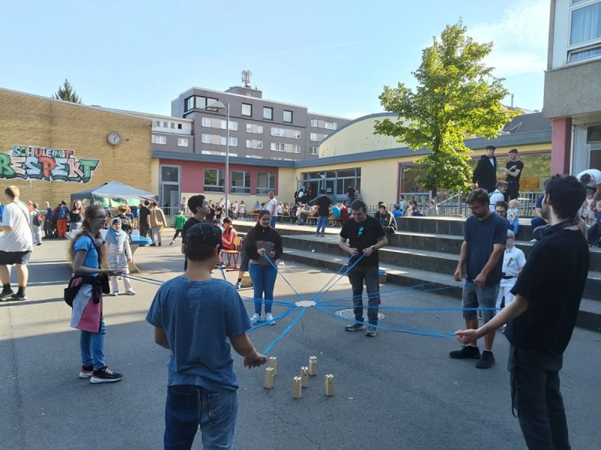Ein weitläufiger Platz vor dem Schulgebäude der LVR-Wilhelm-Körber-Schule. Überall sind Kinder, Jugendliche und Erwachsene zu sehen. In der Mitte spielen neun Schüler*innen ein Spiel mit Seilen und Holzblöcken.